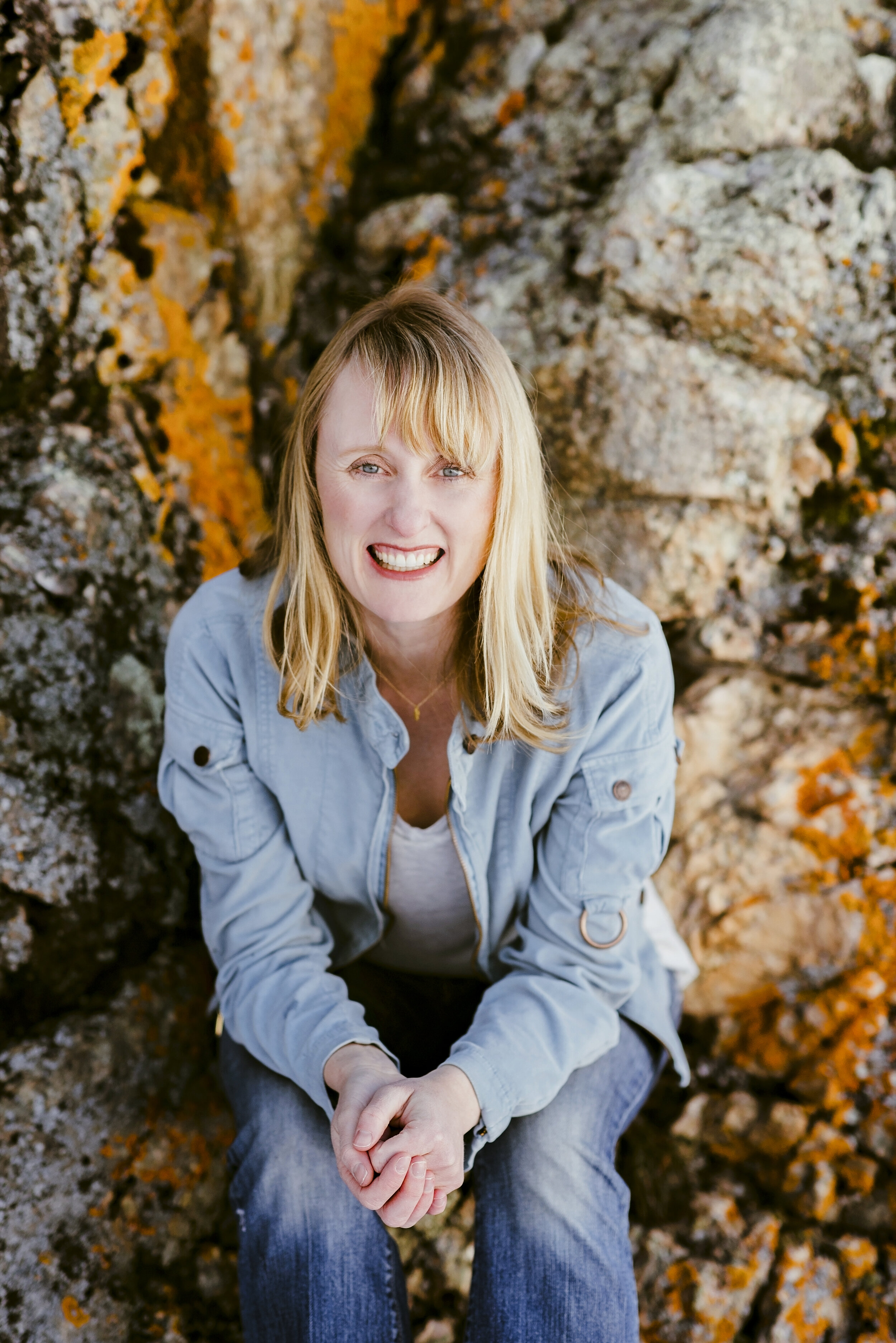 A smiling woman with blonde hair and blue eyes, wearing a light blue denim shirt, sitting on rocks with orange and yellow lichen.