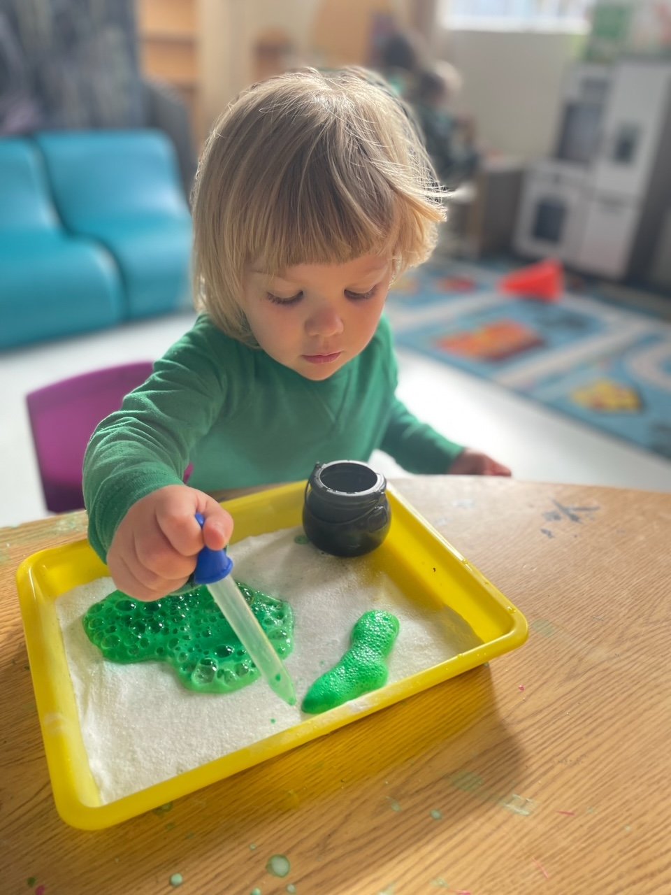 A young child with blonde hair wearing a green shirt is playing with a science kit filled with green slime at a wooden table.