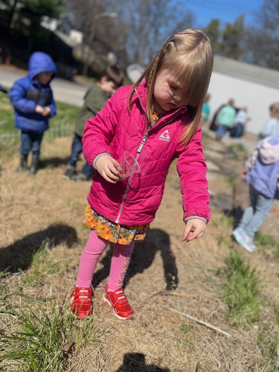 A young girl wearing a pink jacket, floral dress, pink leggings, and red shoes is outdoors, holding a small clear container and examining something on the ground. Other children and adults are in the background, some wearing colorful jackets, on a su