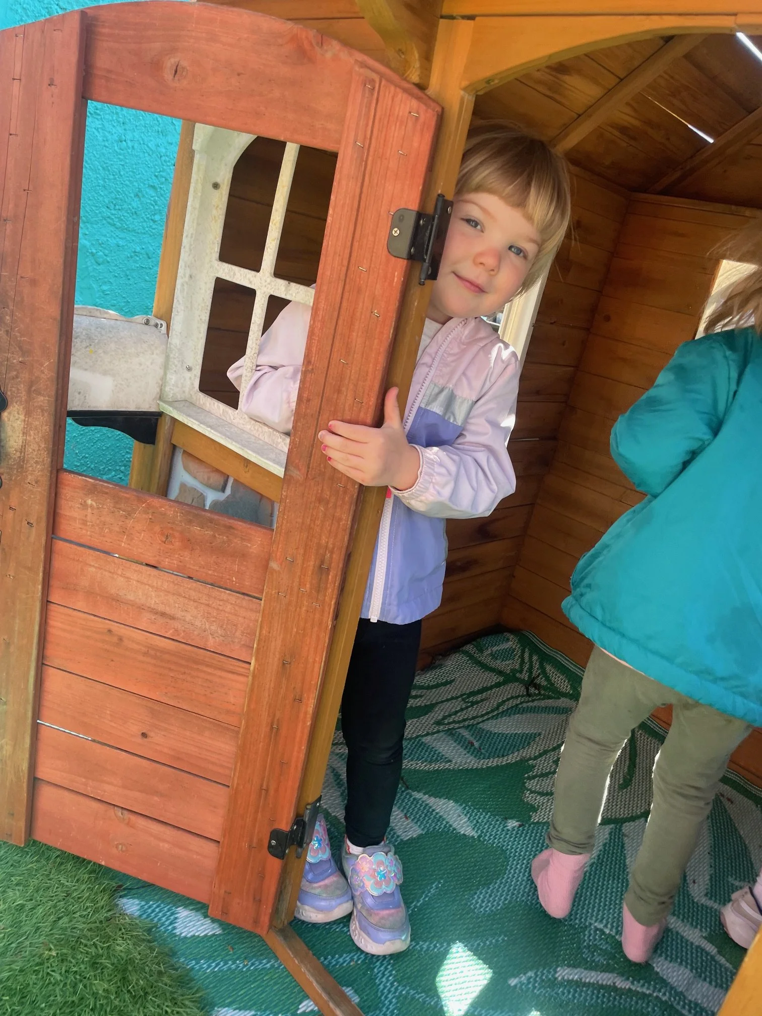 A young girl with light brown hair peeking out from a small wooden playhouse door, smiling and partially inside. She is wearing a light pink and purple jacket, black pants, and colorful sneakers. Another child in a teal jacket and olive green pants i