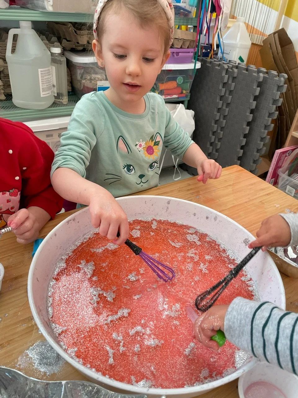 A young girl with a floral headband and green shirt with a cat face, stirring a pink and white mixture with a whisk at a wooden table, with colorful storage containers and foam mats in the background.