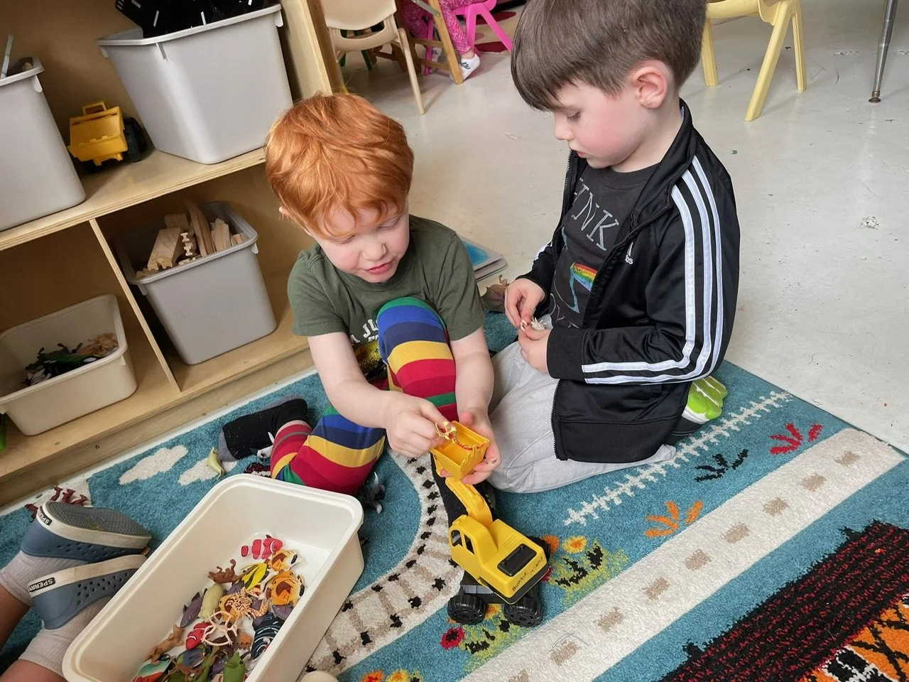 Two young boys playing with toys on a colorful rug in a playroom. One boy has red hair and is wearing rainbow-striped pants, holding a yellow toy excavator. The other boy has brown hair and is wearing a black jacket with a rainbow flag patch, kneeling beside him.