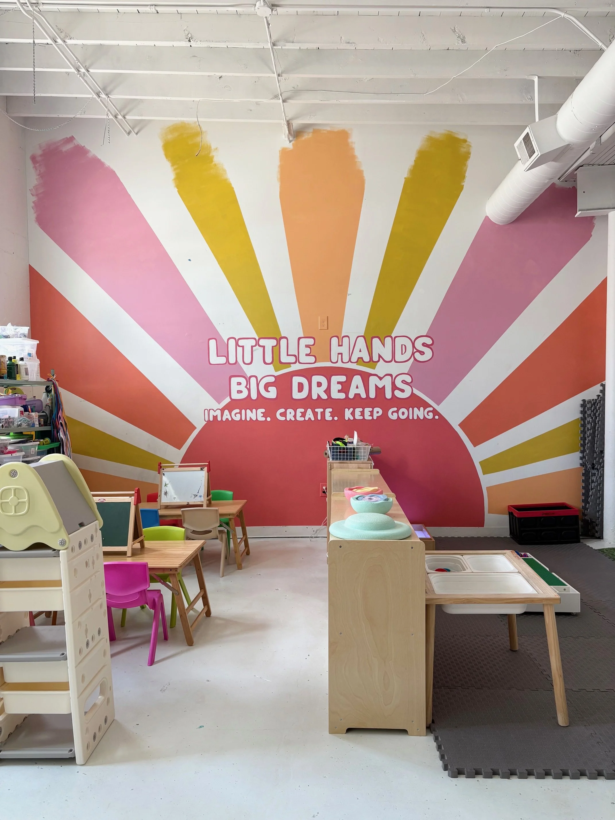 Colorful playroom with a mural on the wall reading "Little Hands Big Dreams" and "Imagine. Create. Keep Going." There are small tables, chairs, plush cushions, and toys, with a soft mat on the floor.