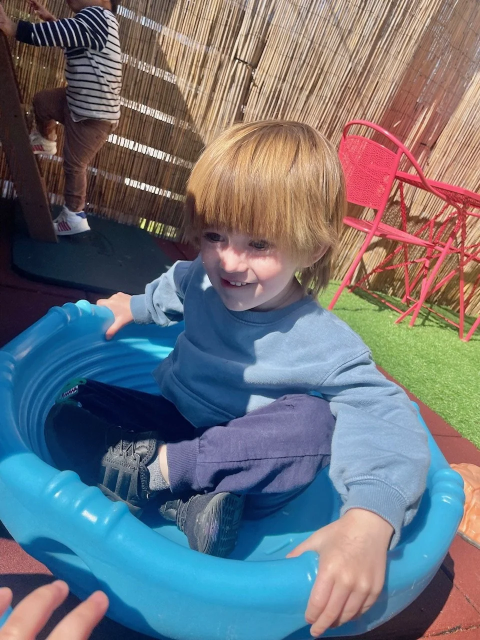 A young boy with red hair is sitting in a small blue plastic swimming pool outdoors, smiling and playing. There is a pink chair and a green artificial turf mat in the background, with a bamboo fence surrounding the area.