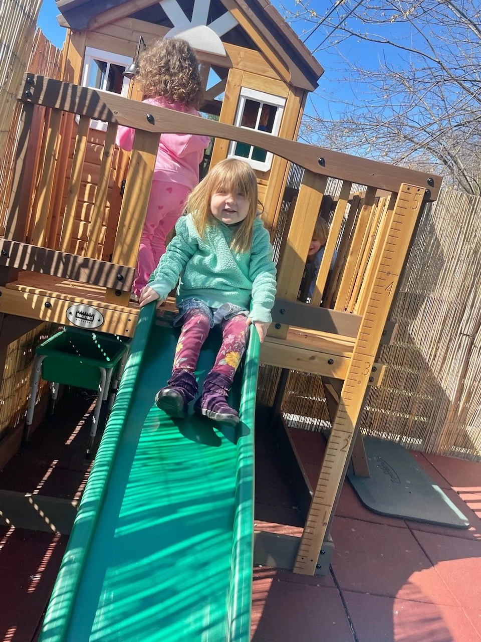 Two young girls on a playhouse slide outdoors on a sunny day, with a wooden playhouse and a bamboo fence in the background.