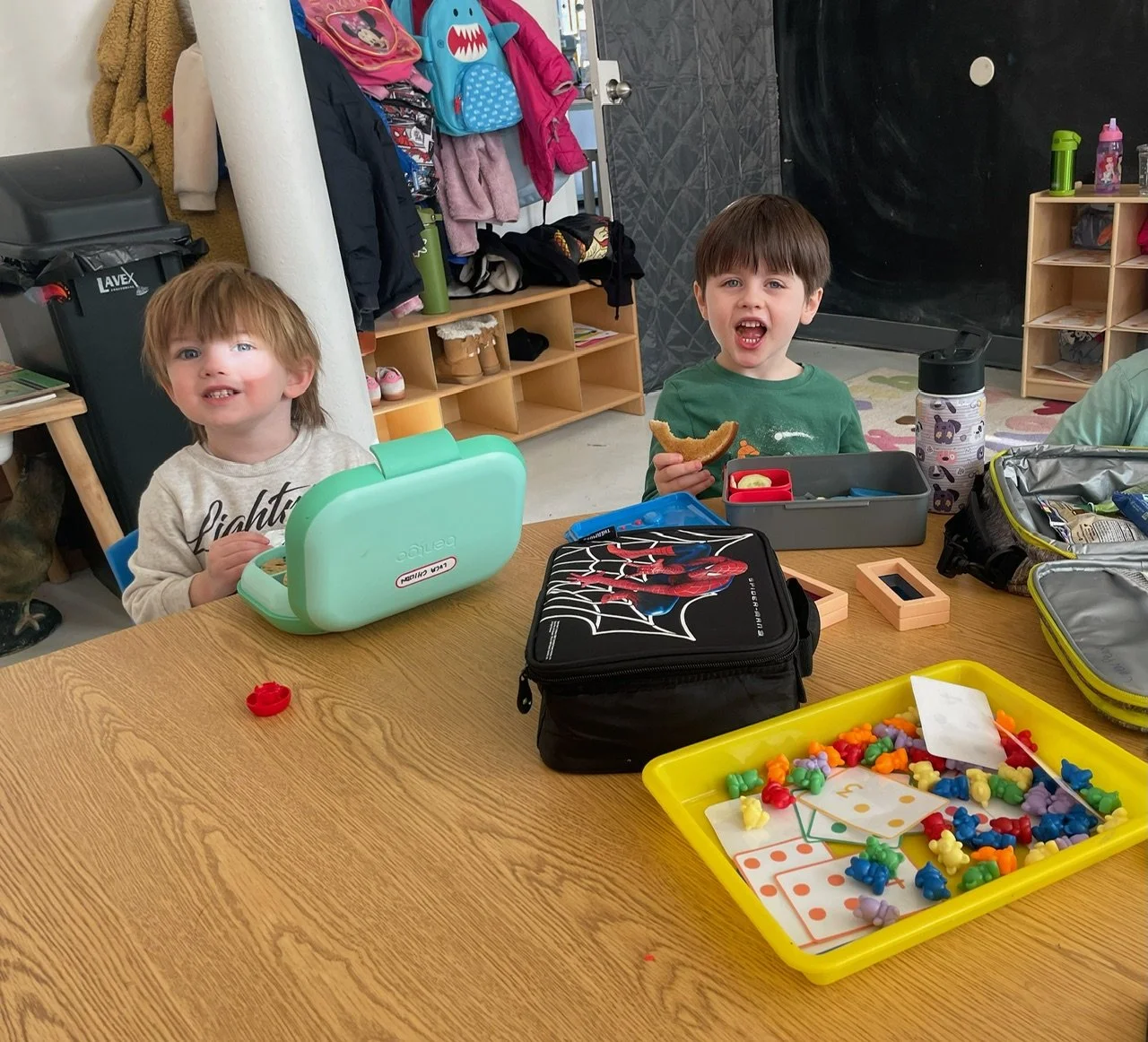 Two young children sitting at a table during snack time in a classroom, with lunchboxes, toys, and classroom supplies on the table.