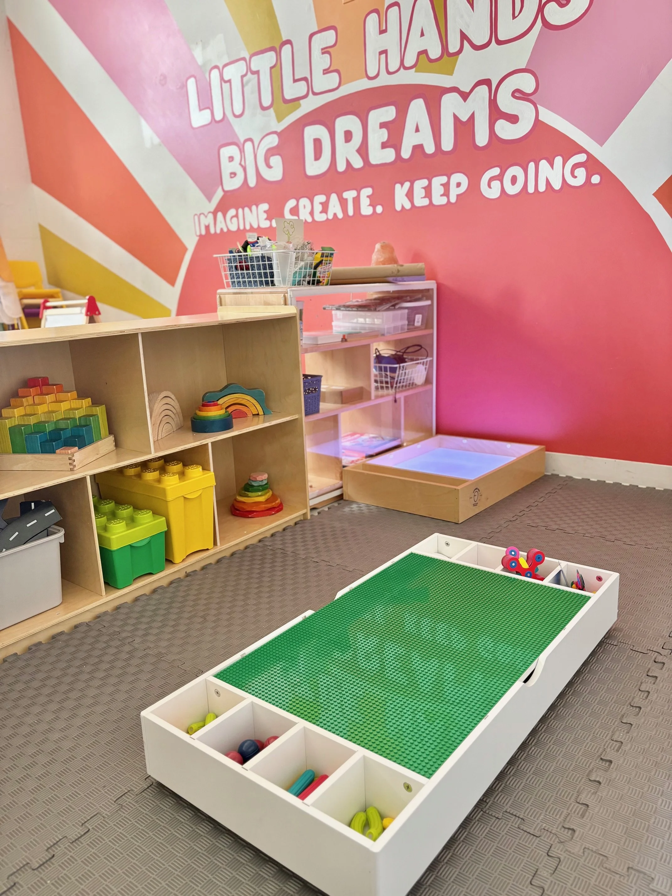 Children's play area in a colorful indoor space with a large pink wall, a green Lego table, shelves with various toys and blocks, and a mural with the words "Little Hands Big Dreams" and "Imagine. Create. Keep Going."