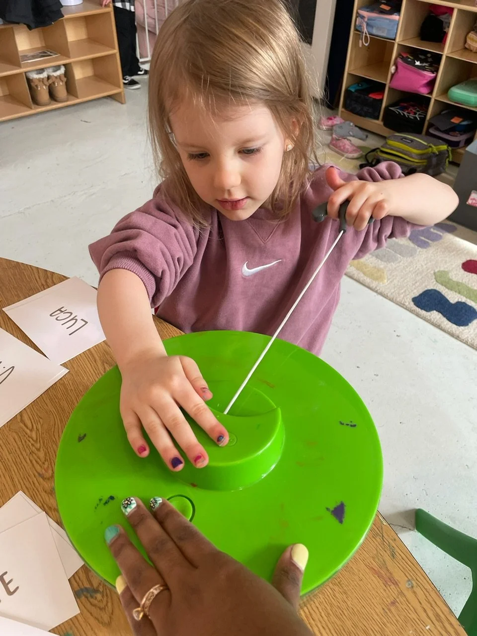 A young girl with blonde hair uses a green spinning wheel toy to make yarn in a classroom setting.