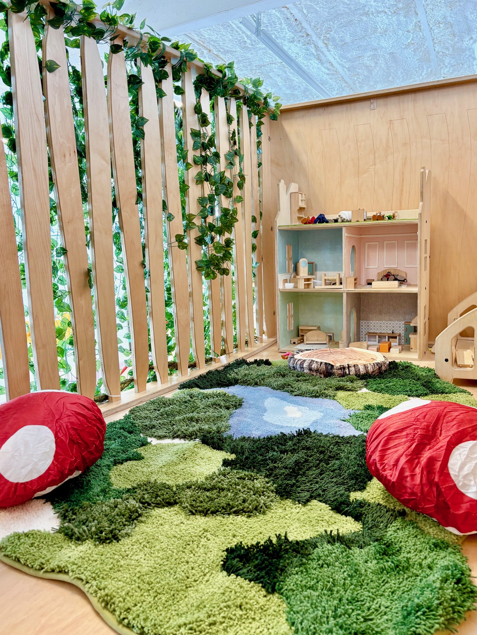 A children's playroom with a green, nature-themed carpet, red paper lanterns, a wooden dollhouse, and a wall of greenery behind a wooden lattice.