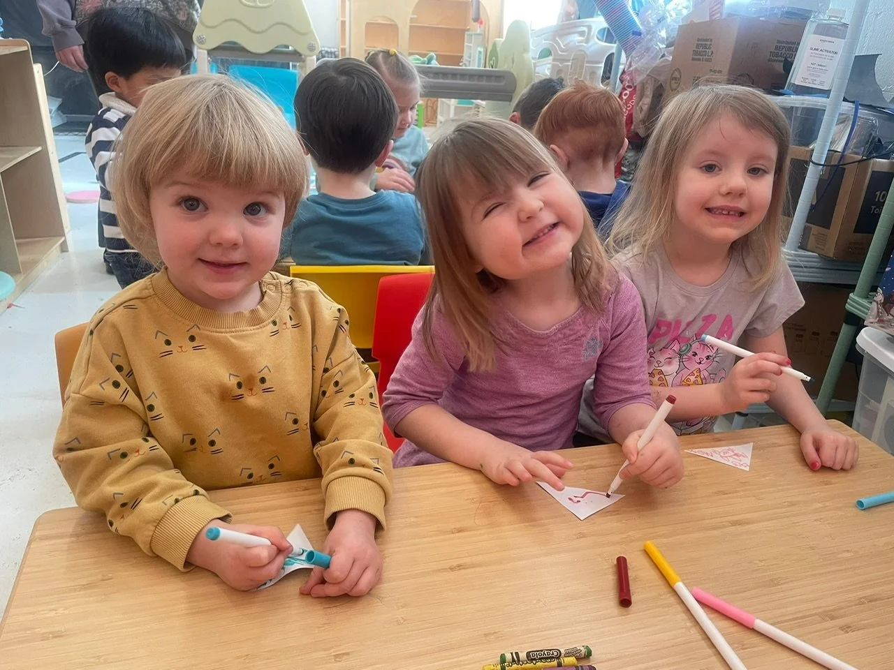 Three young girls smiling and drawing with markers at a table in a classroom, with other children and shelves in the background.