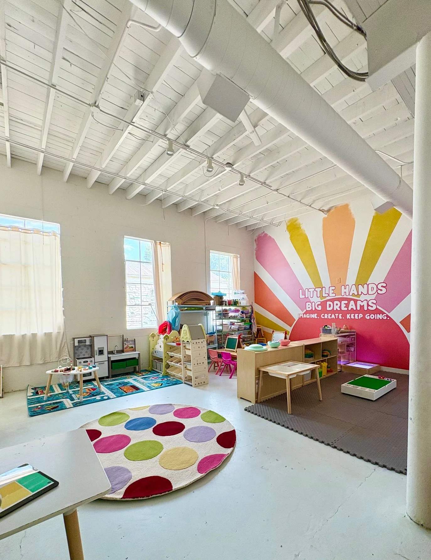 Colorful children's playroom with toys, tables, and a patterned rug. A bright pink wall with yellow sunburst design and inspirational quote: 'Little Hands Big Dreams. Imagine, Create, Keep Going.'