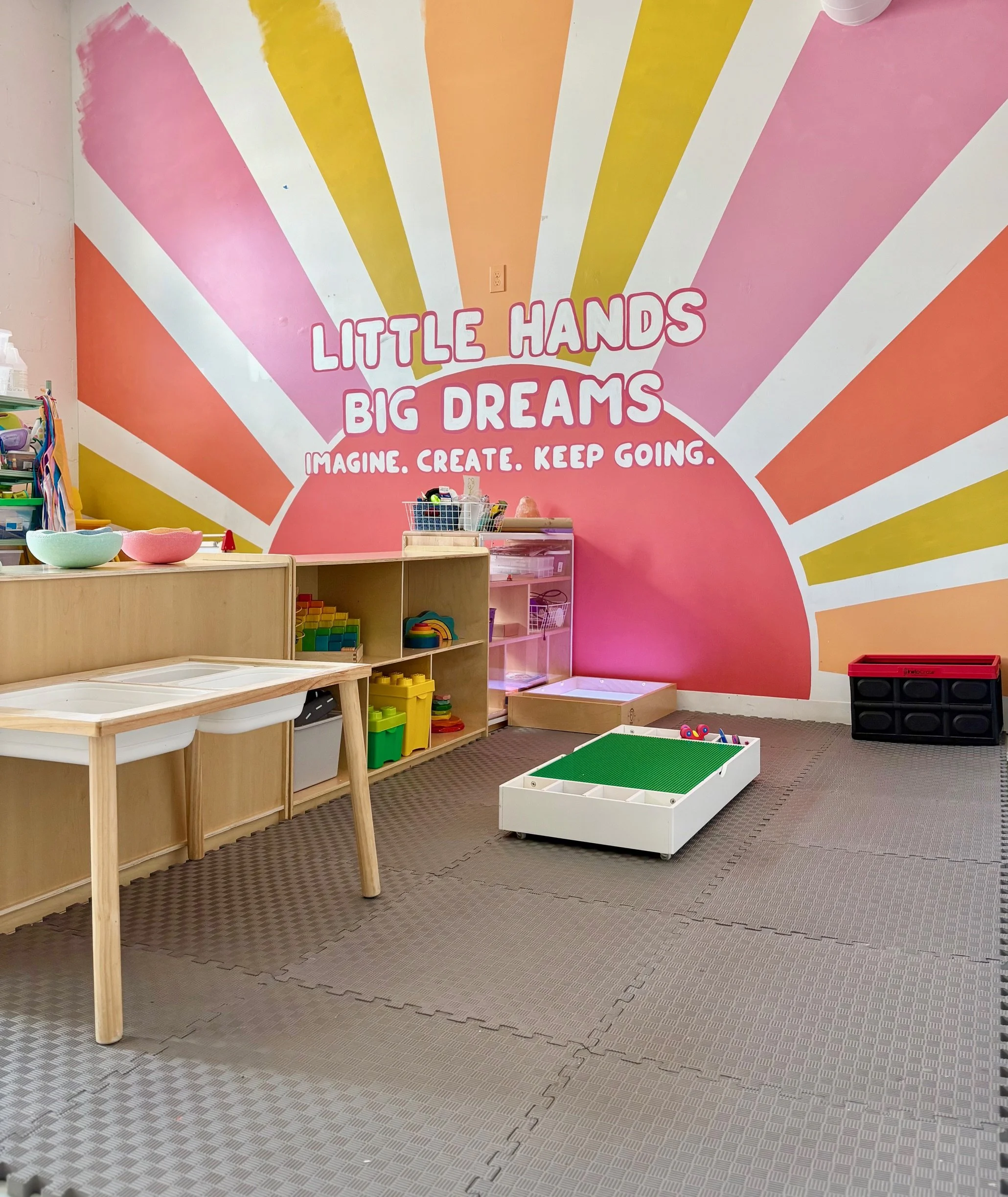 Colorful children's playroom with a pink, yellow, orange, and peach wall mural that says, "Little Hands Big Dreams. Imagine. Create. Keep Going." There are wooden and plastic shelves with toys, bowls on a table, and a green-boarded sandbox on a rubbe