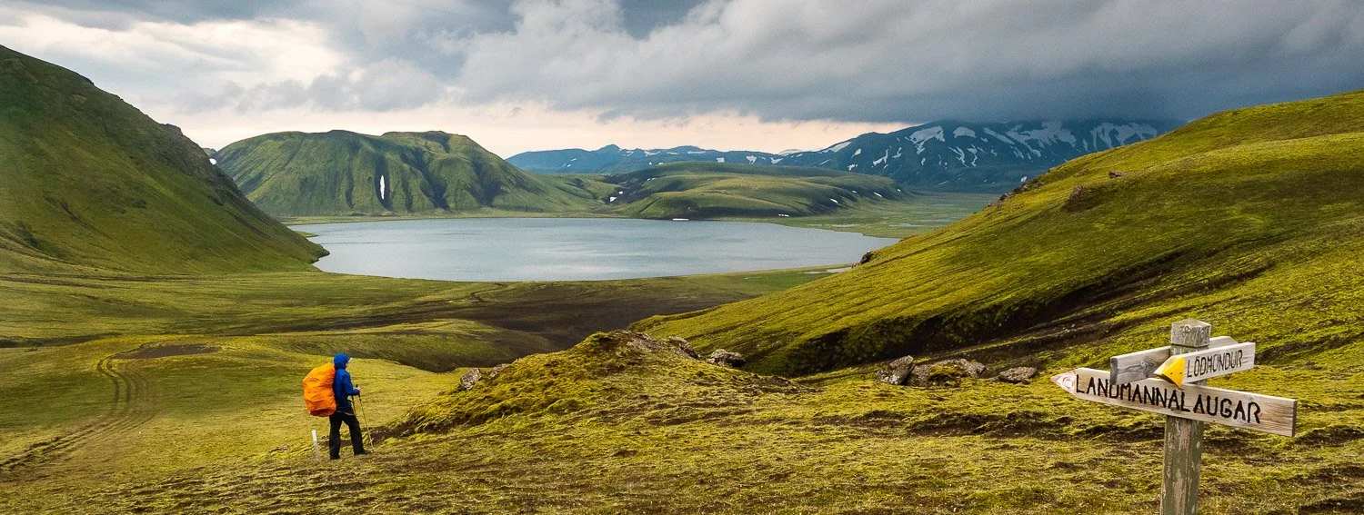 hiker on flat grassy area admiring view of small lake and surrounding mountains with patchy snow.