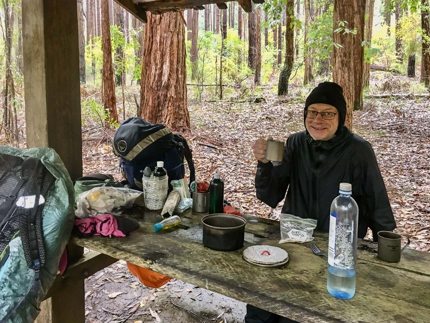 hiker sitting at picinic bench under small shelter in rain gear having a cup of soup