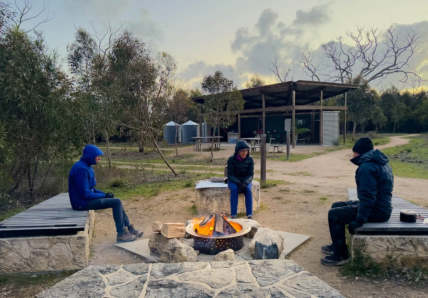 three hikers rugged up sitting around a fire pit at camp site