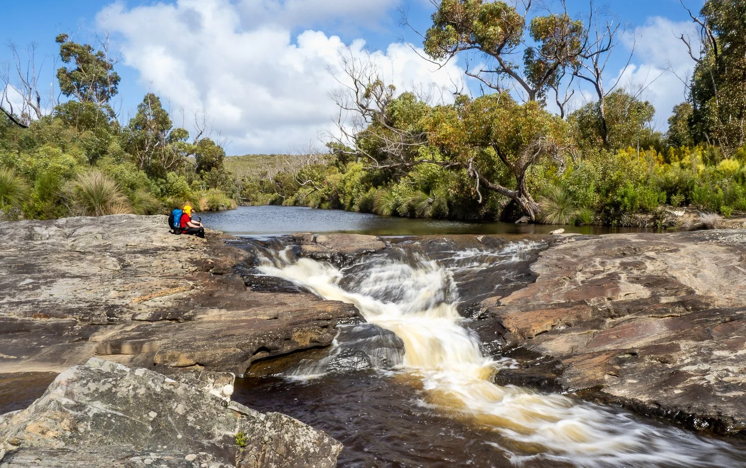 hiker with red shirt and blue backpack sitting at head of cascade with long still pool of water behind