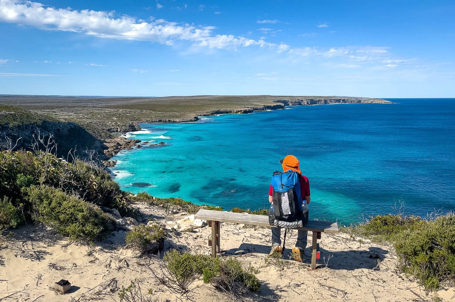 hiker sitting on seat near cliff top under sunny sky looking down at seals in calm sea below
