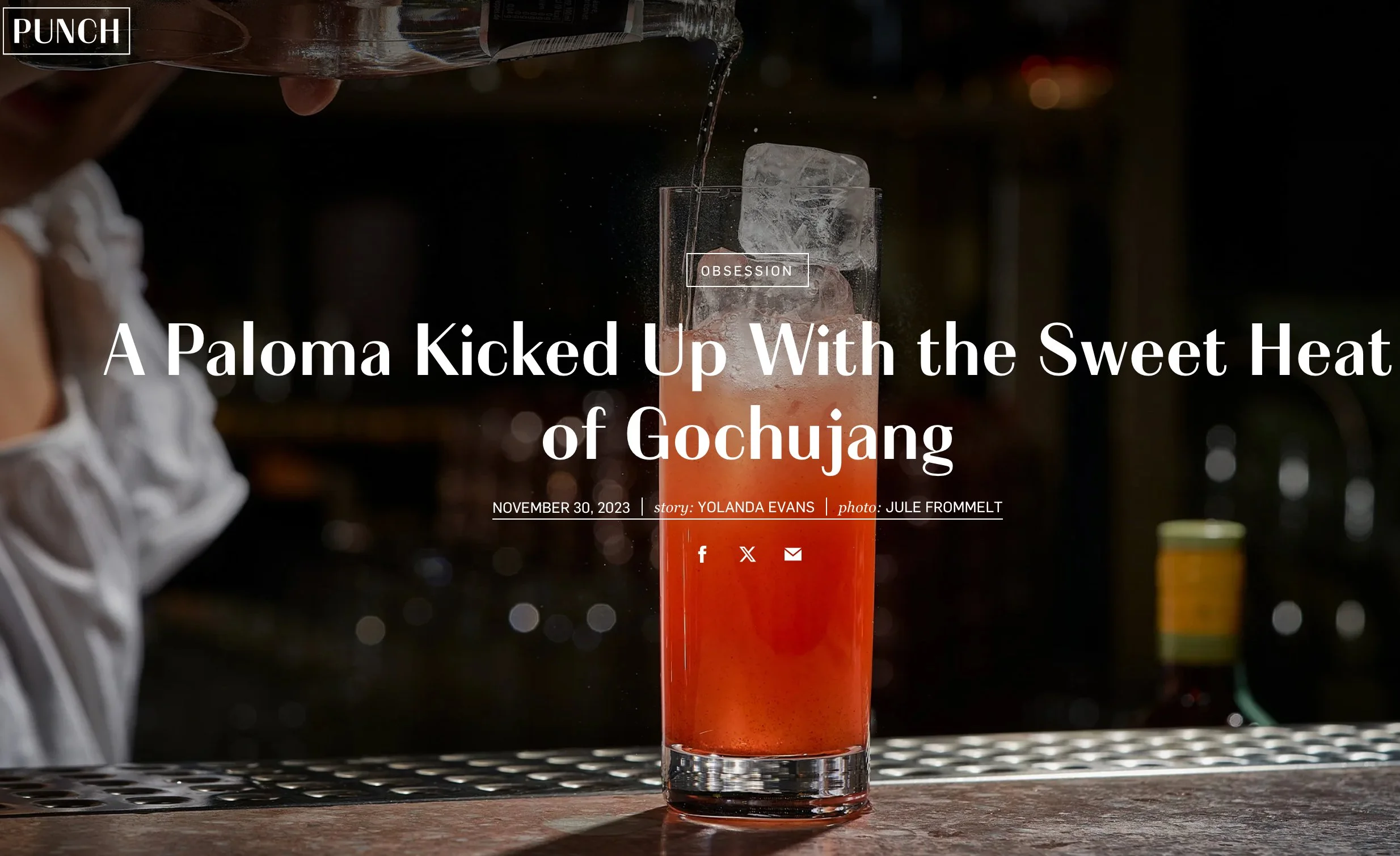 A tall glass filled with red-colored beverage and ice cubes, being poured with liquid from above, on a bar counter with a blurred background.
