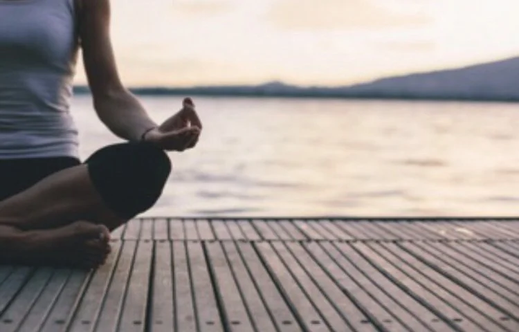A woman in a black top and beige wide-leg pants standing in a field of tall, golden grass on a partly cloudy day, holding the edges of her pants.