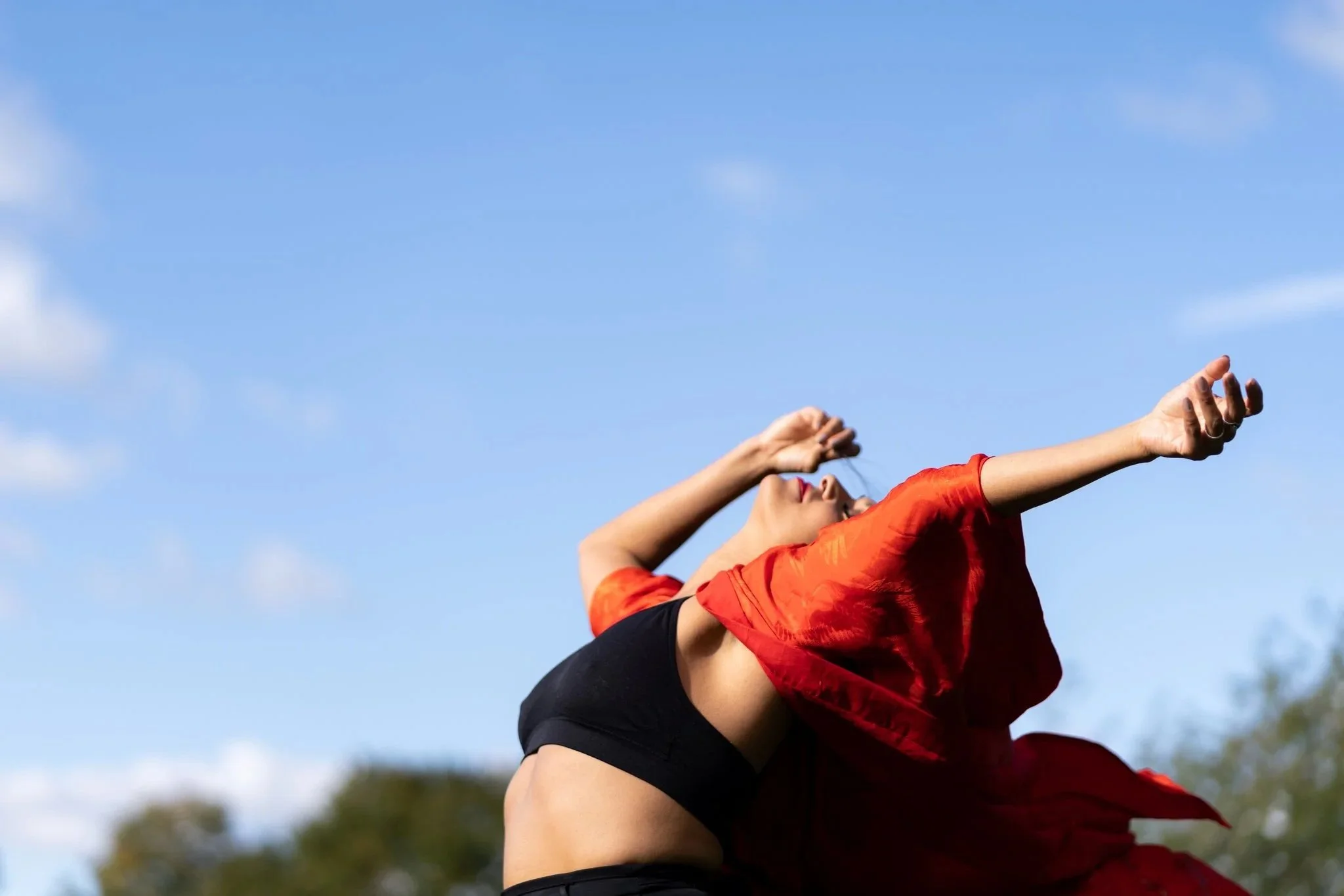 A woman dancing outdoors under a blue sky with scattered clouds, wearing a black top and red flowing fabric.