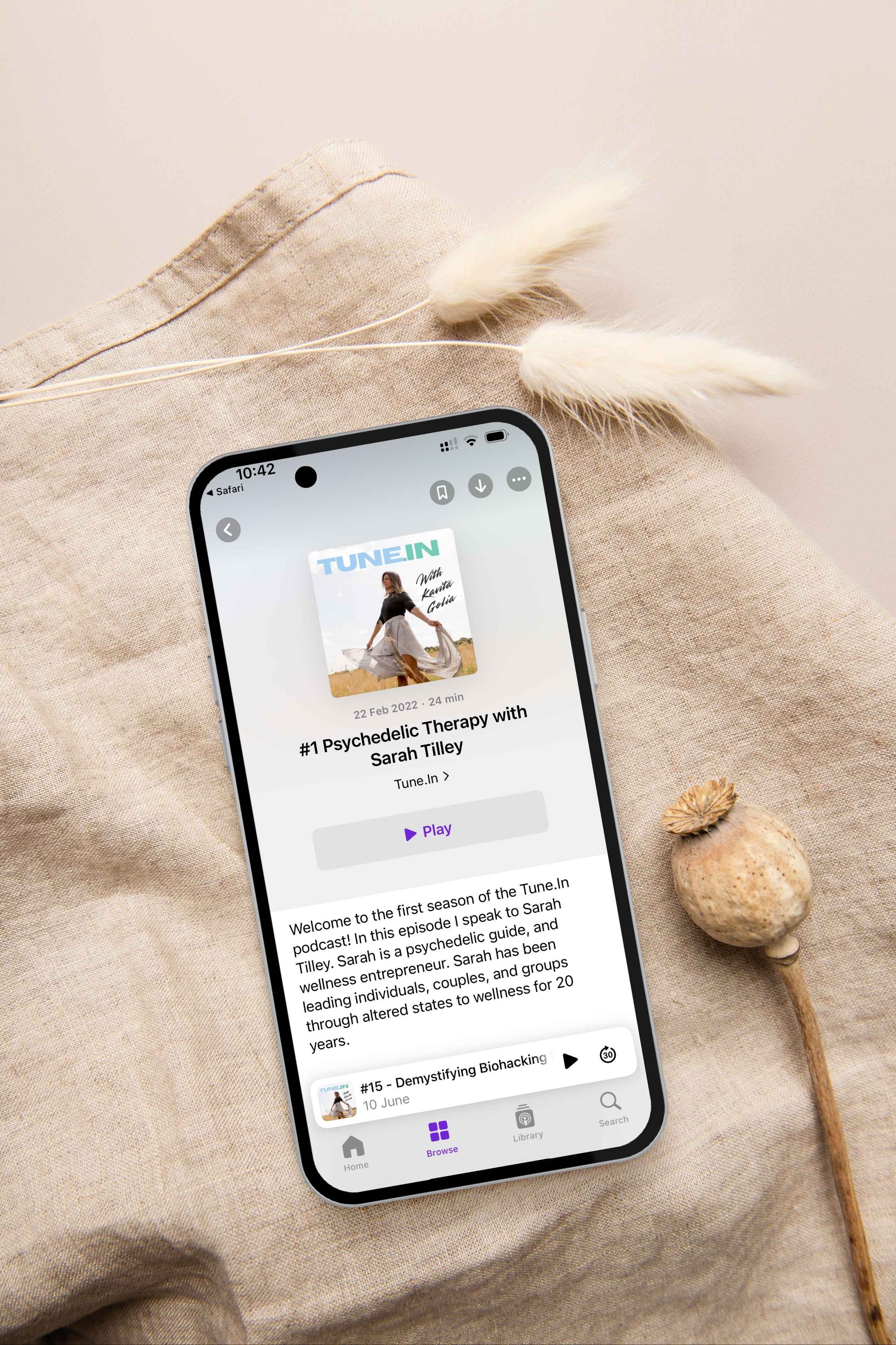 Smartphone displaying a podcast titled '#1 Psychedelic Therapy with Sarah Tilley' on a beige fabric surface, with dried flowers nearby.