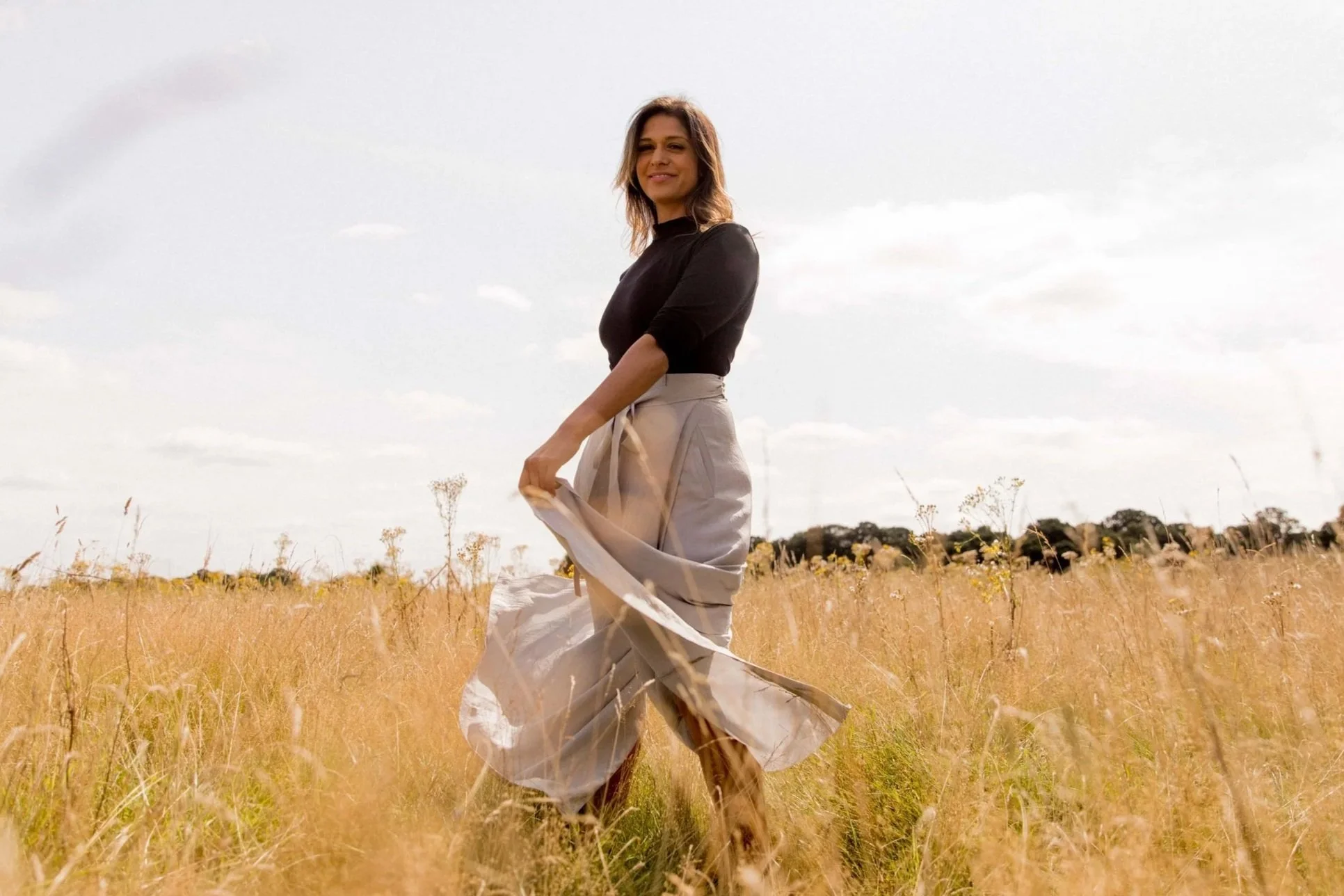 A woman sitting in a field of tall, golden grass, wearing a white dress, with her head tilted back and eyes closed, enjoying the sunlight on a clear day with trees in the background.