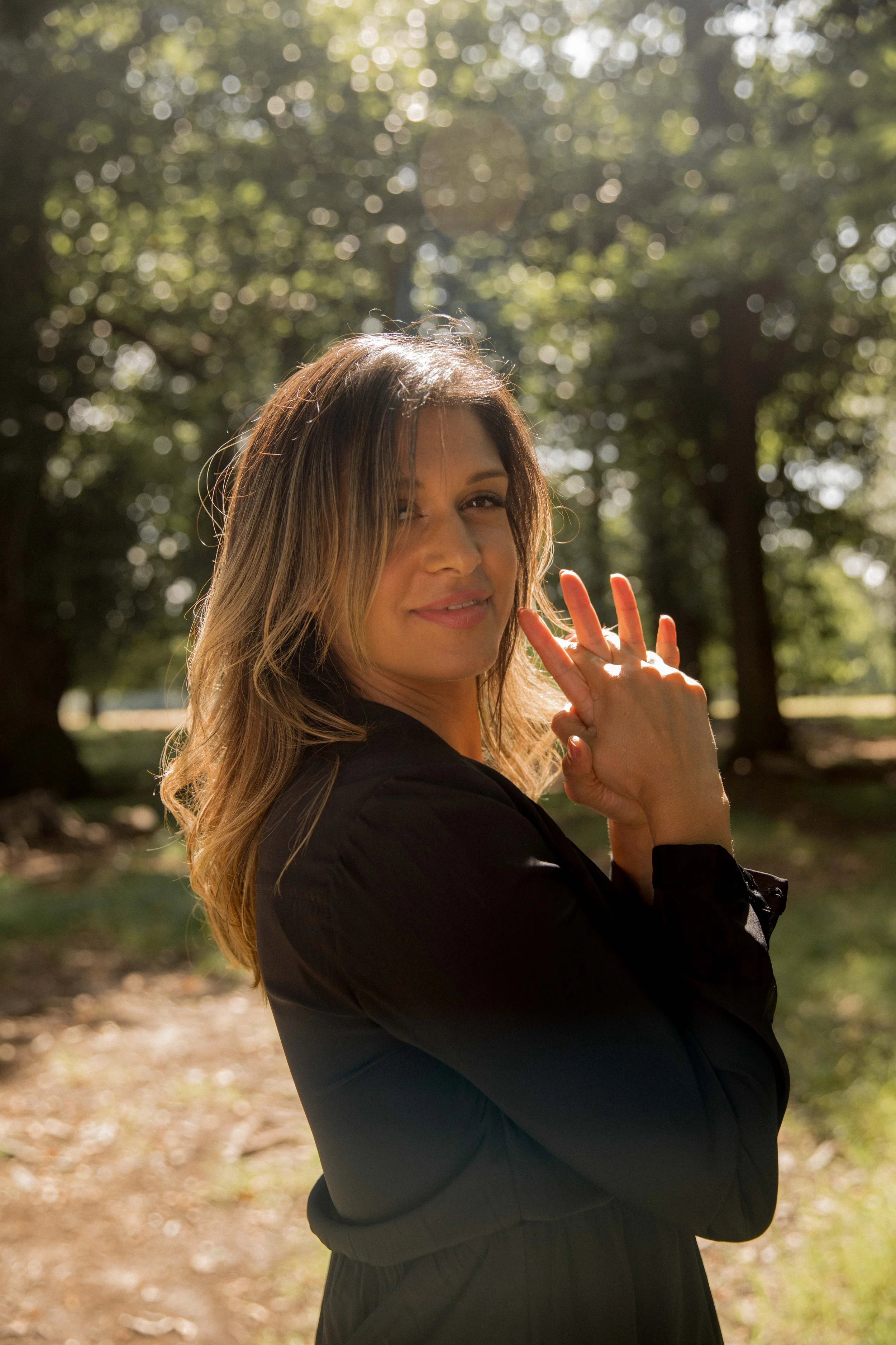 A woman with wavy brown hair winking backwards and making a peace sign in a park with sunlight filtering through trees.