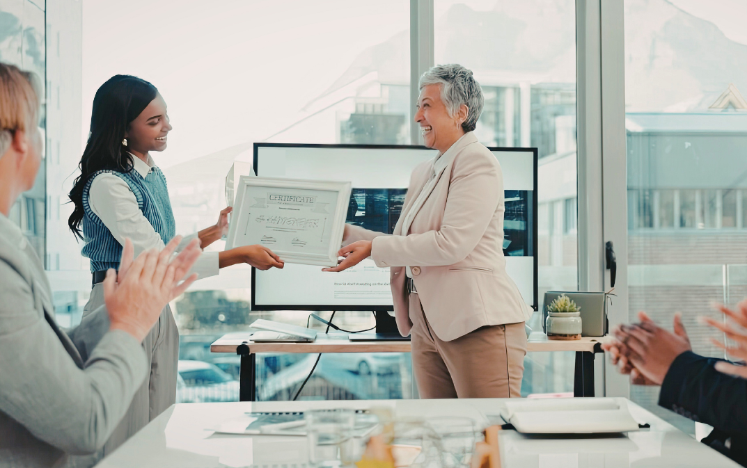 A woman receives a certificate in an office while her colleagues applaud her.