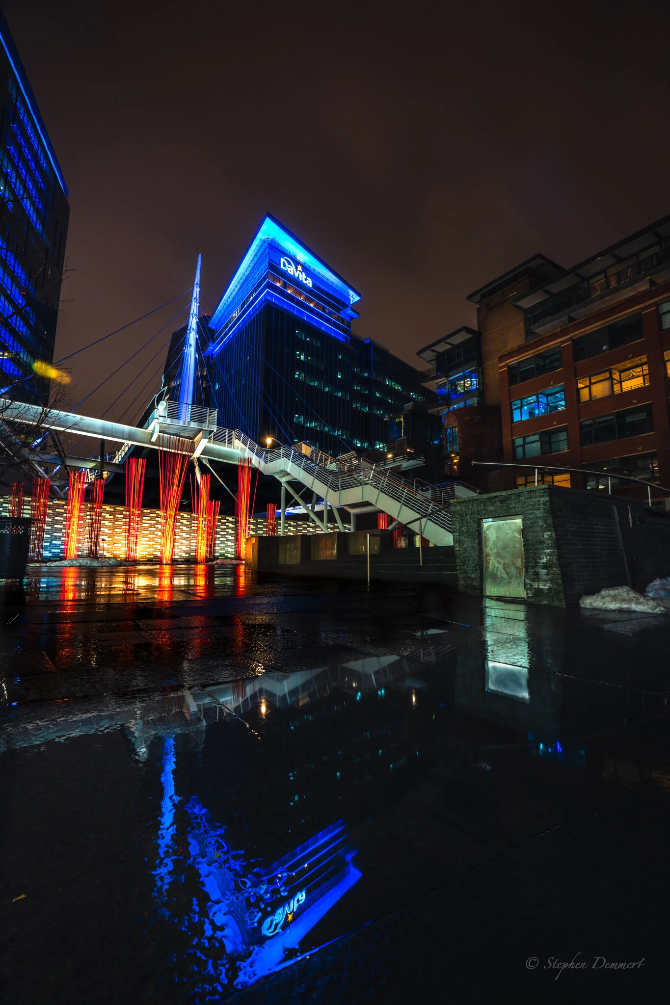 Nighttime cityscape with illuminated modern buildings, colorful lights reflecting on wet ground, and a visible sign reading 'DaVita' on a tall building.