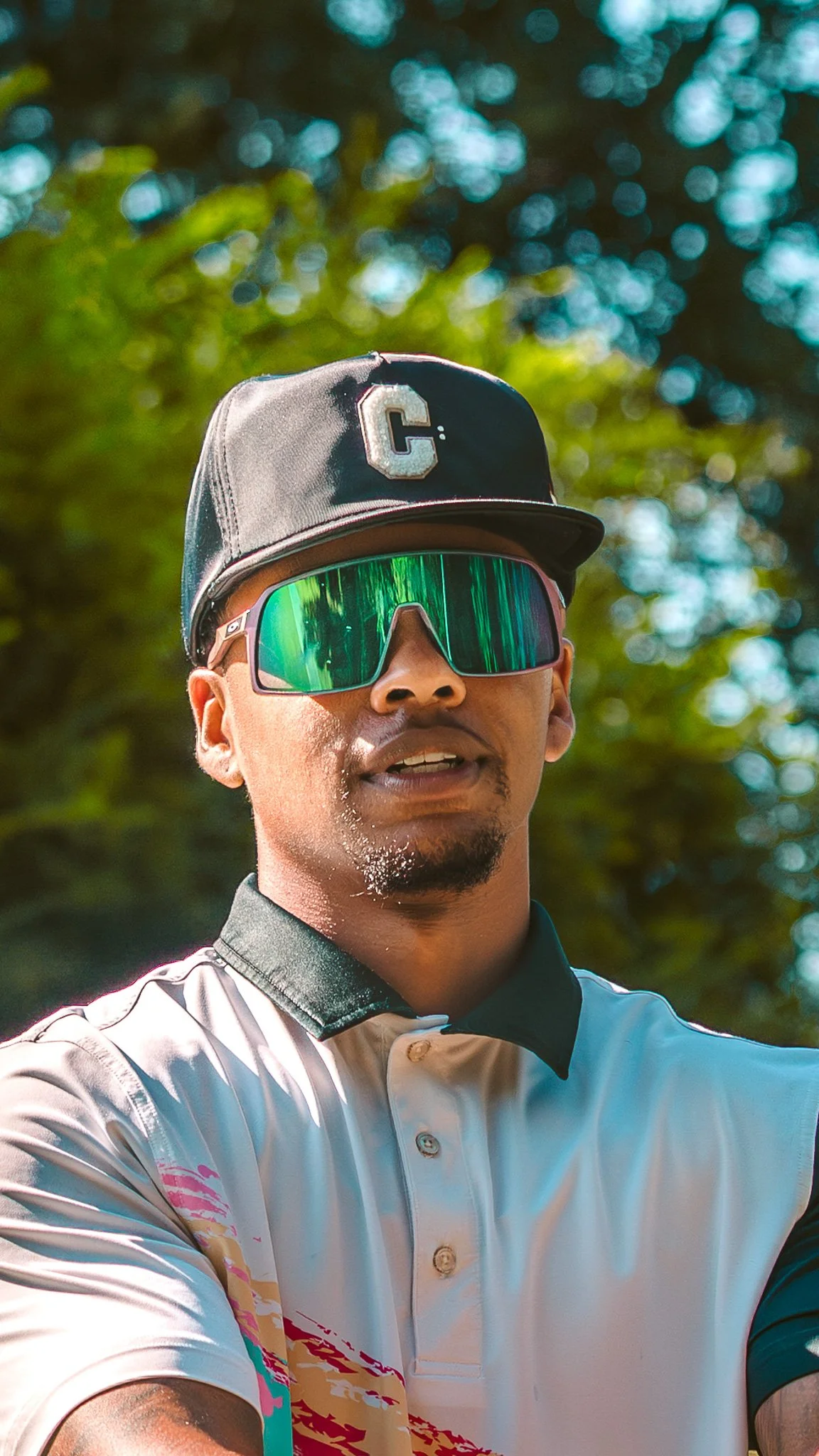A man wearing a black cap with a 'C' on it, reflective sports sunglasses, and a light-colored polo shirt with a black collar outdoors with green trees in the background.