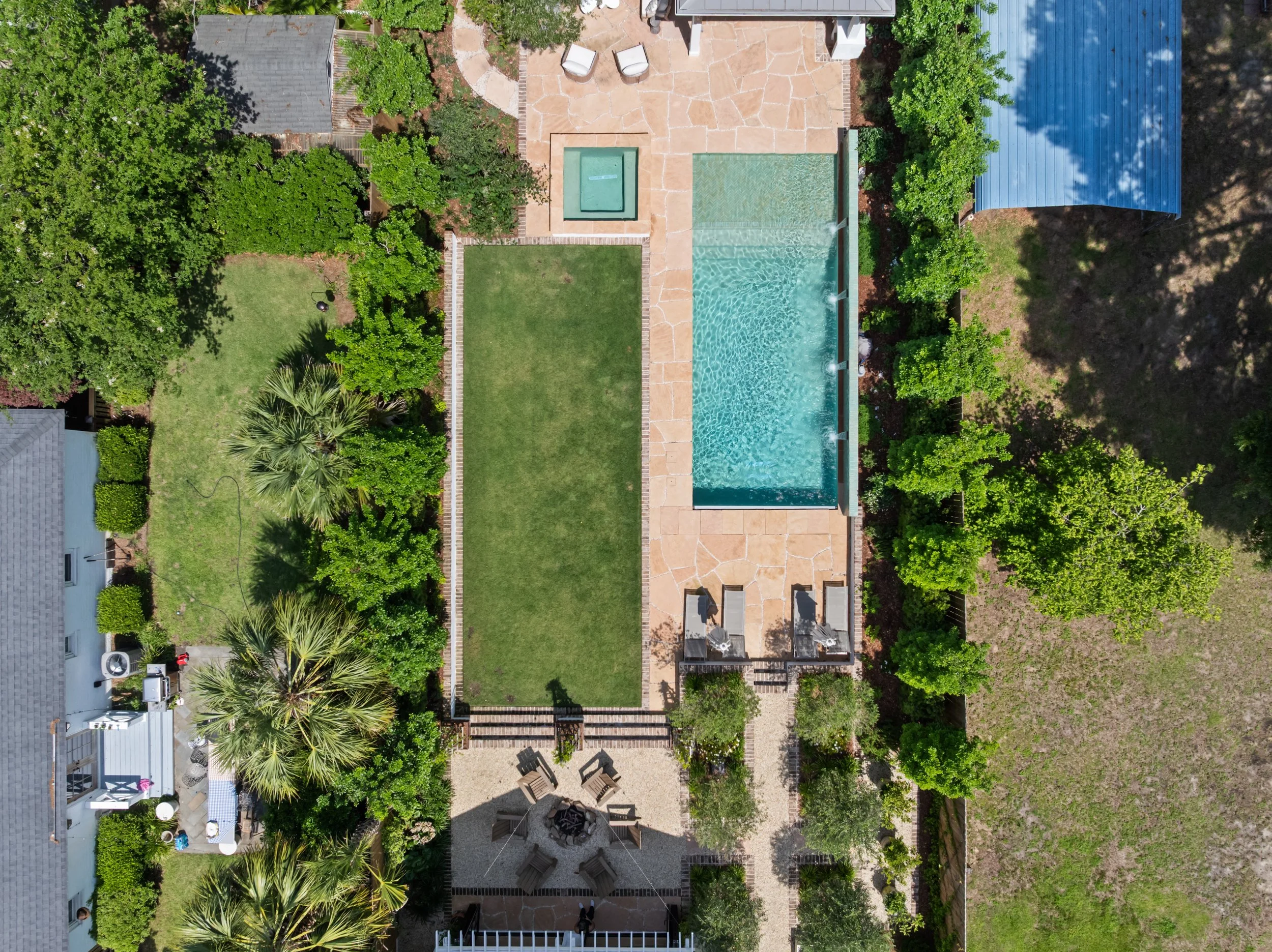 Aerial view of a backyard with a swimming pool, hot tub, grassy lawn, outdoor seating area with chairs and a fire pit, and surrounded by green trees and shrubs.