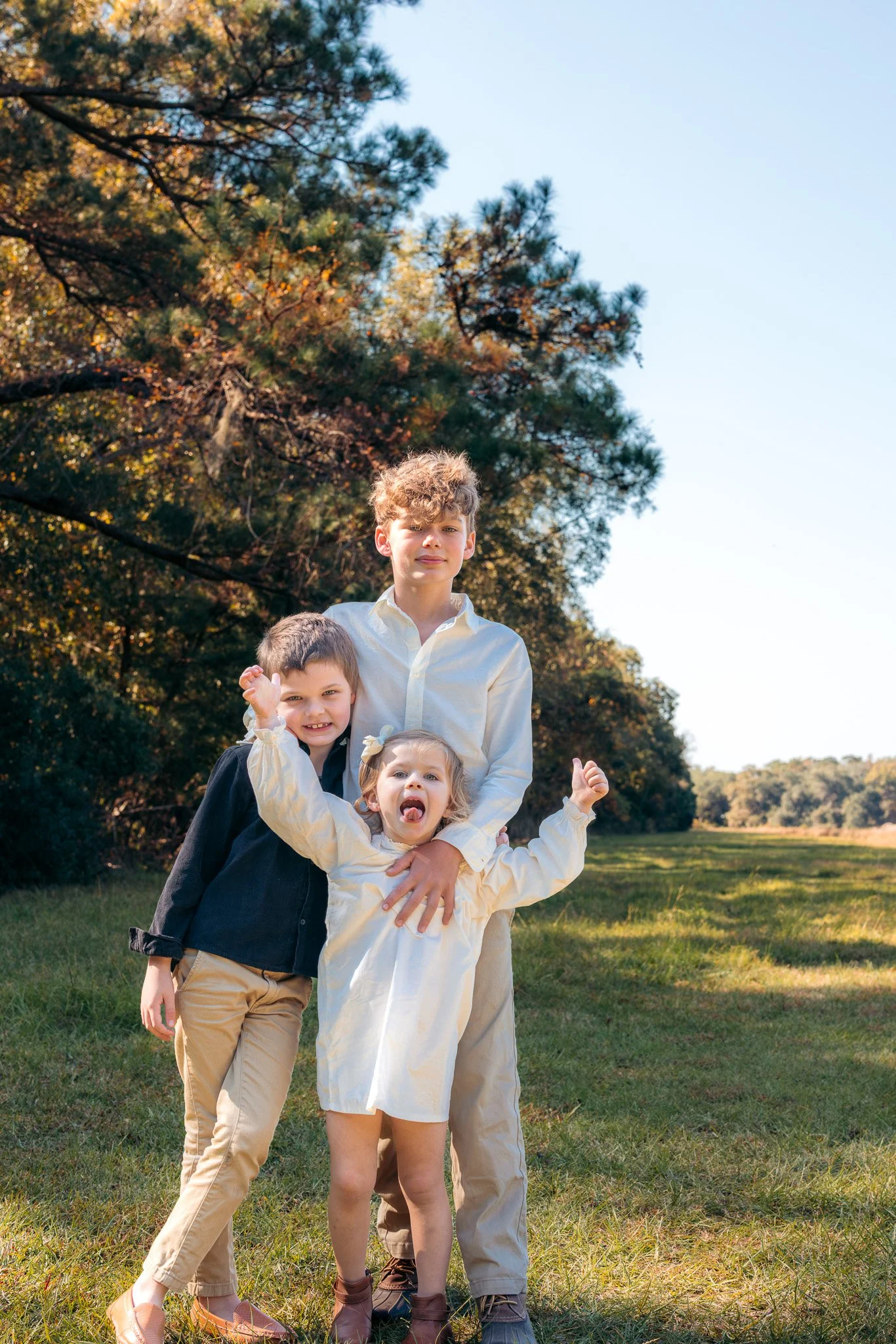 Three children standing outdoors on a sunny day, with trees in the background. The oldest boy is in the center, wearing a white shirt. To his left is a girl with a bow in her hair, sticking her tongue out and holding her arms up. To his right is a bo