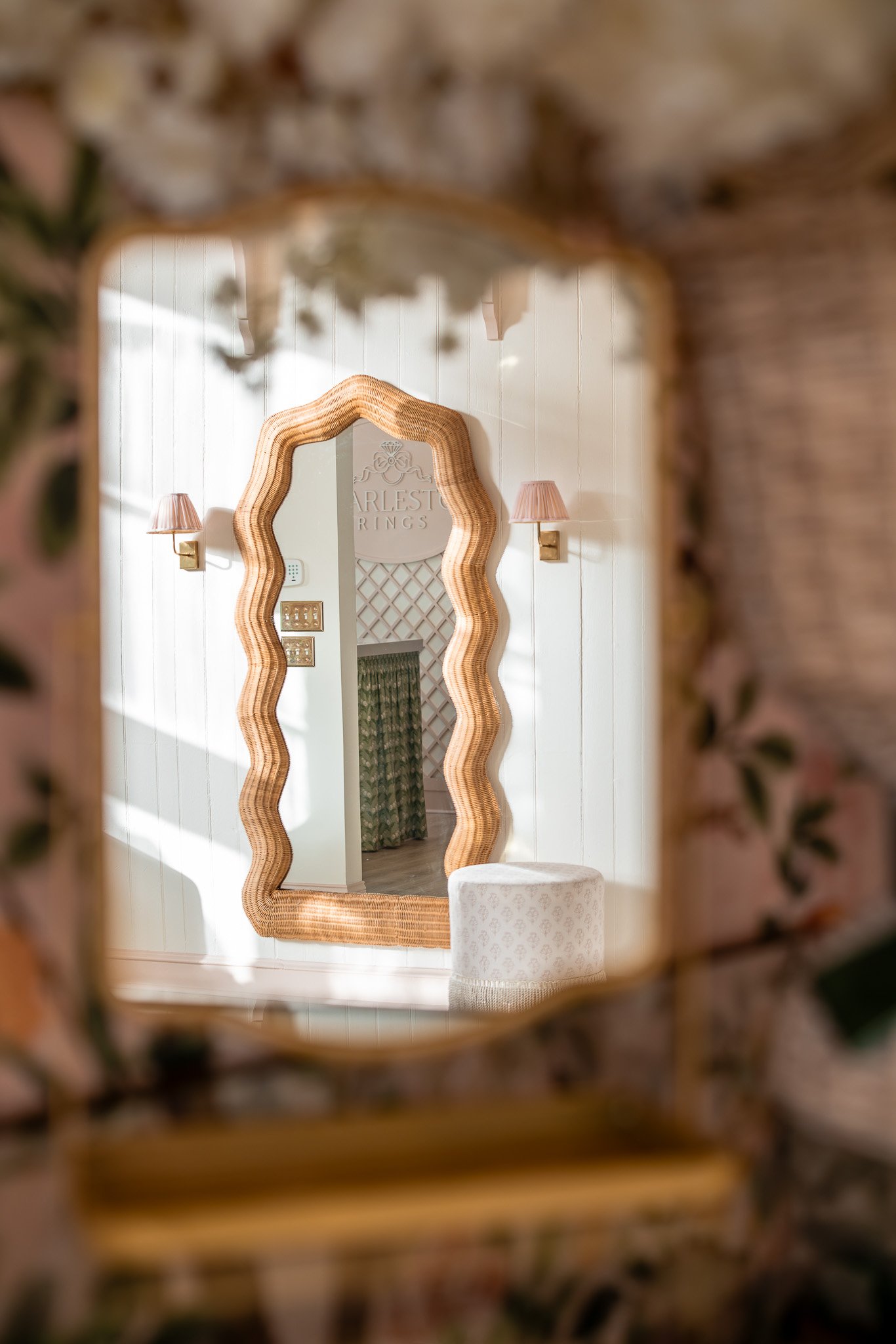 A decorative mirror with a wavy wooden frame reflected in a mirror, hanging on a white-paneled wall, with small pink lampshades on either side.