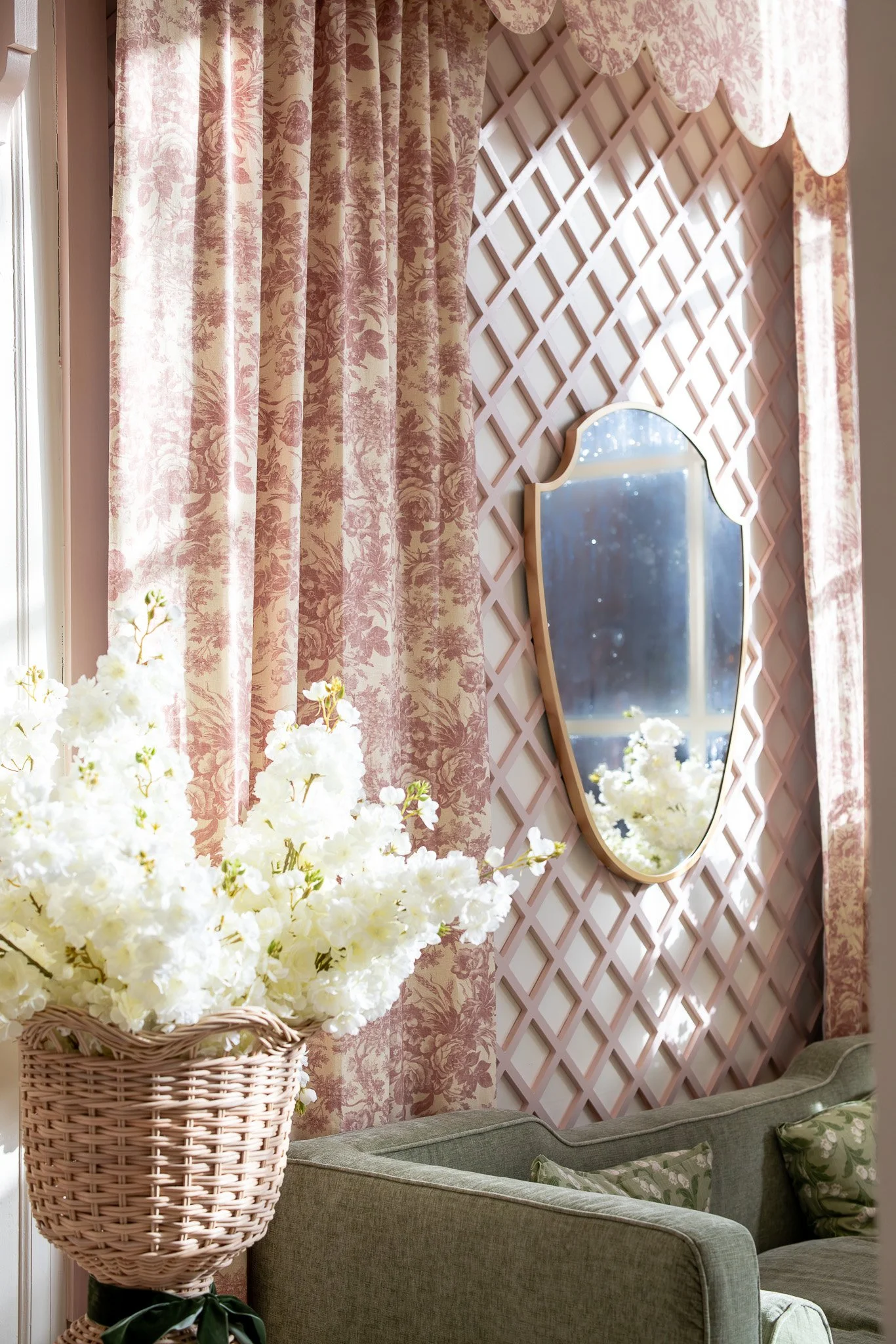 A cozy living room corner with beige floral curtains, a pink lattice accent wall with a decorative mirror, a bouquet of white flowers in a wicker basket, and a green upholstered sofa with floral pillows.