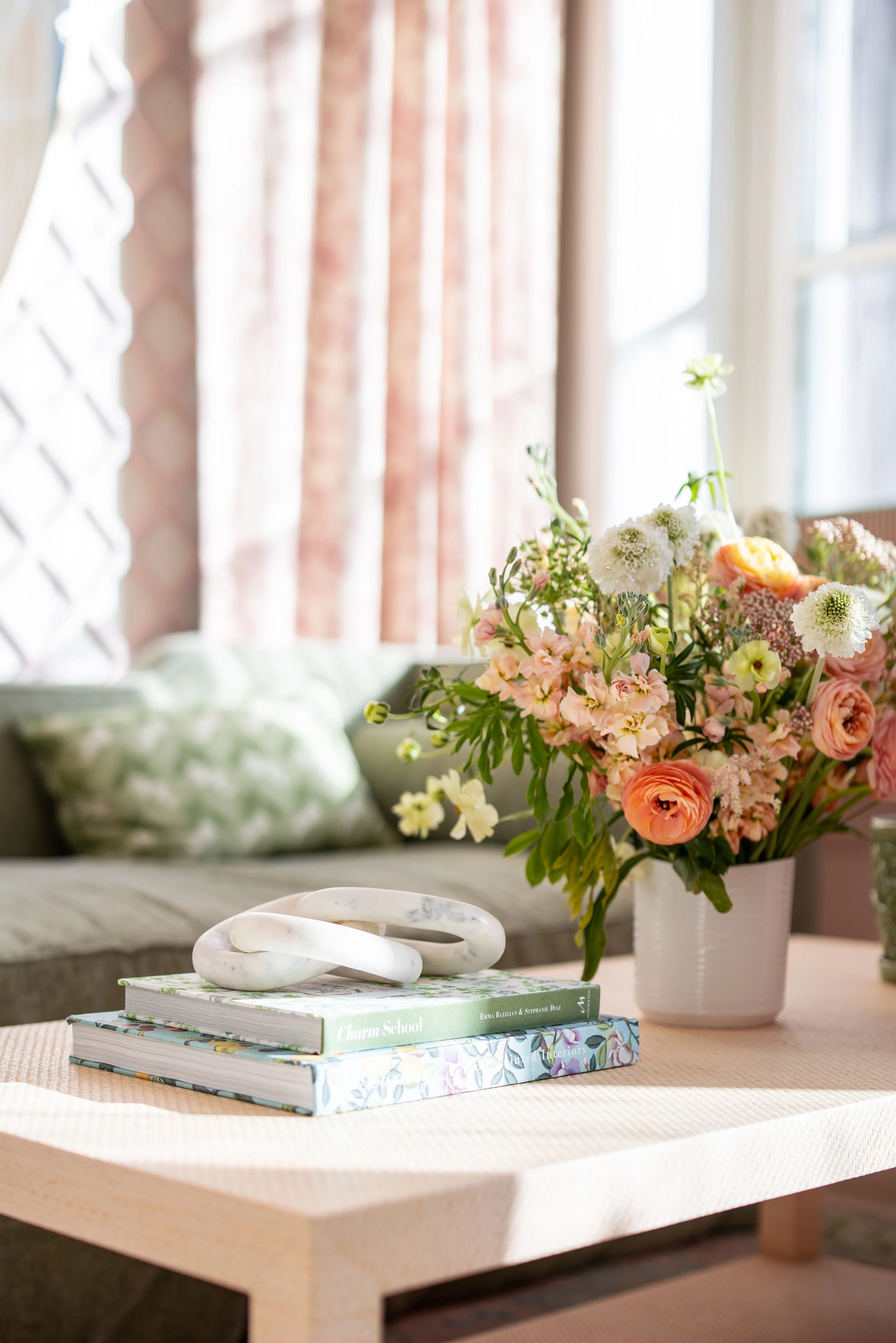 A peach-colored table with a white marble sculpture, three large books, and a white vase filled with pink and white flowers. In the background, there is a couch with green and white pillows and floral curtains next to a large window with sunlight str