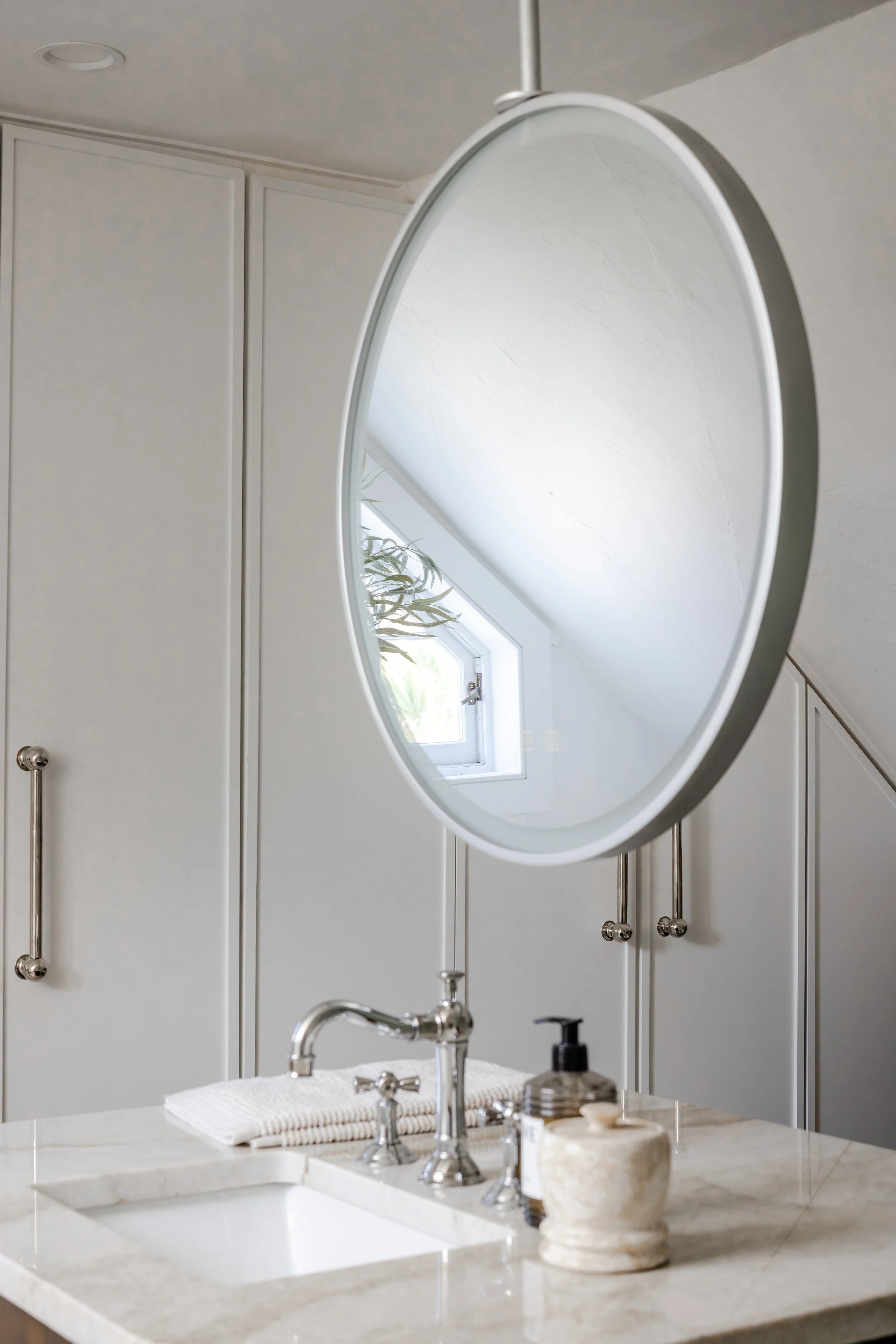A bathroom with a round mirror, a small window, and a sink with toiletries.