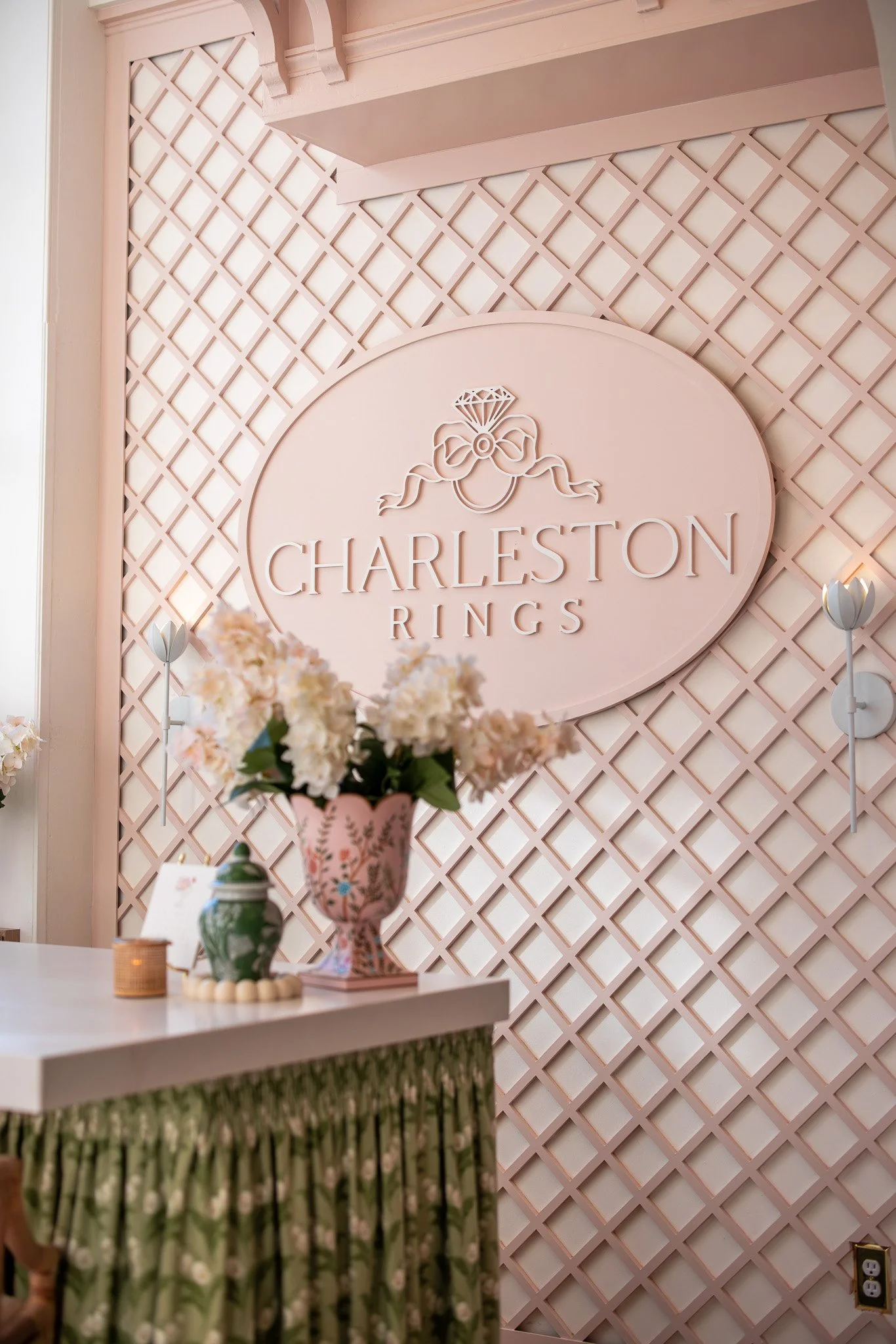The image shows a pink decorative wall with a sign reading 'Charleston Rings,' featuring a diamond and bow design. A table in front has a pink floral vase with white and pink flowers, another smaller green vase, a small candle, and a standing card. T