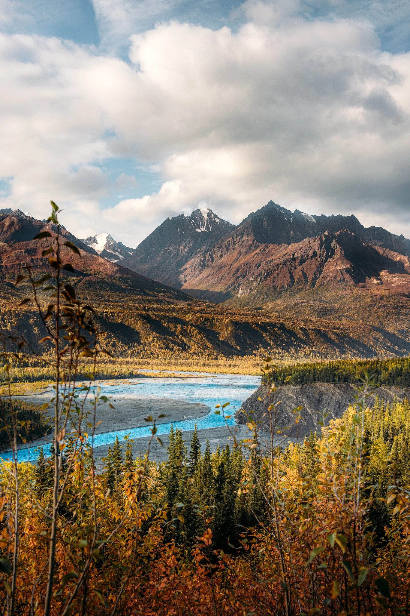 A scenic landscape featuring a river winding through a forested valley with mountains in the background under a partly cloudy sky.