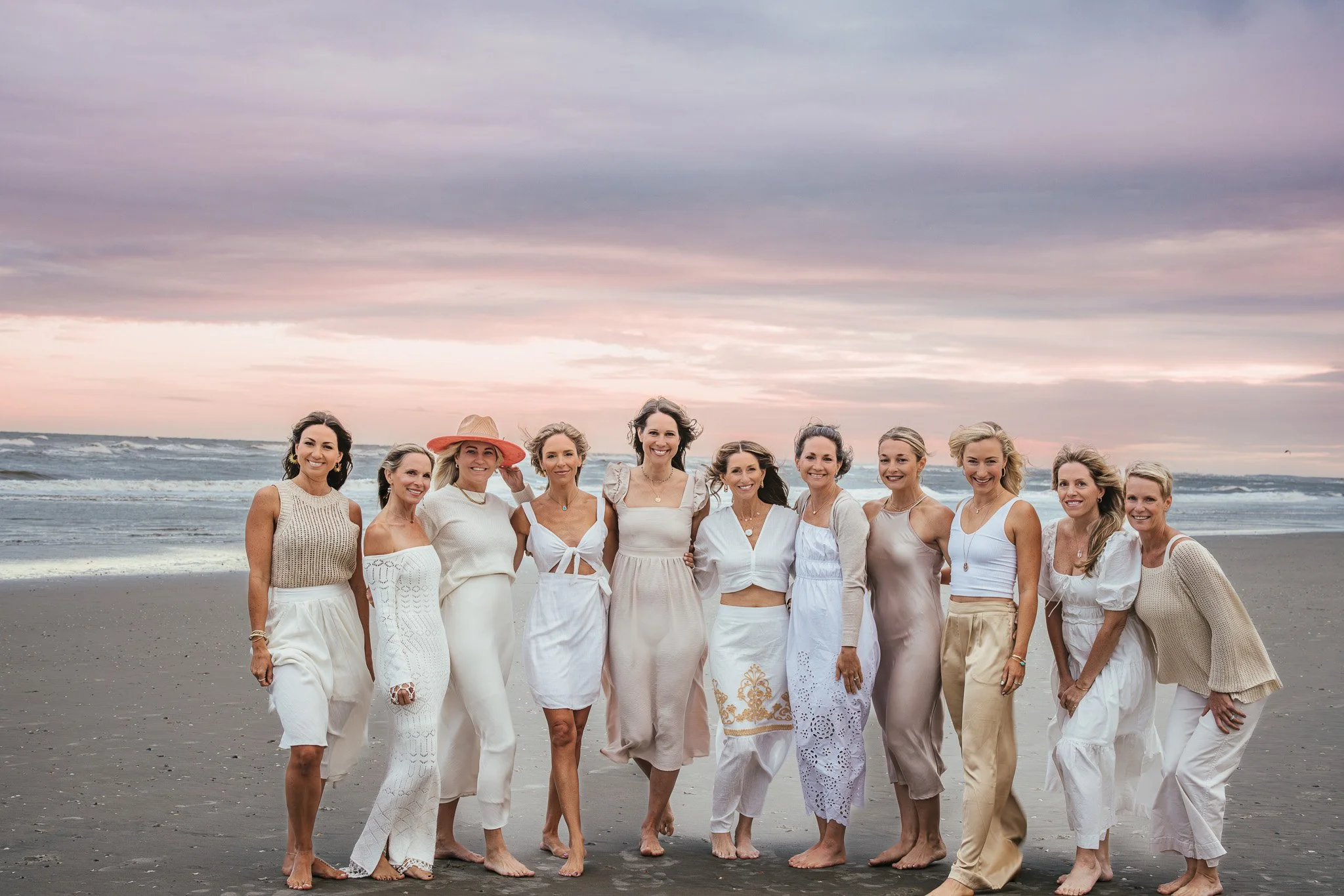 A group of twelve women standing on a beach at sunset, dressed in light-colored clothing.