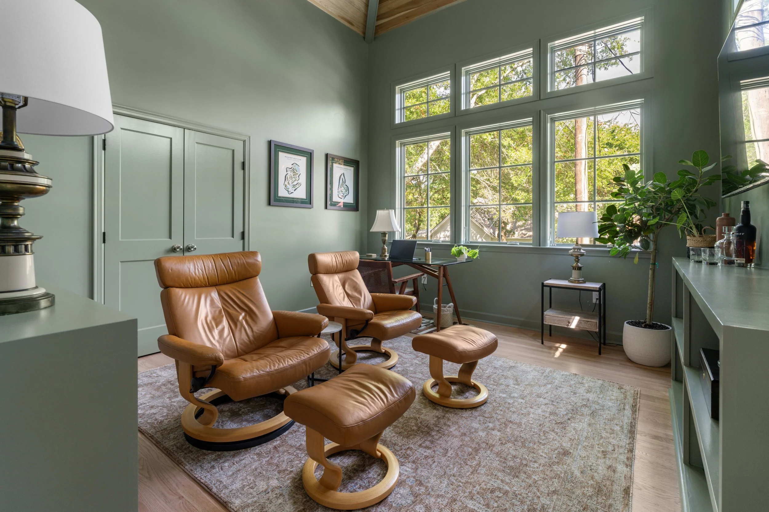 Living room with three tan leather chairs and footstools on a beige rug, green walls, large window with trees outside, side and table lamps, potted plant, and black picture frames on the wall.