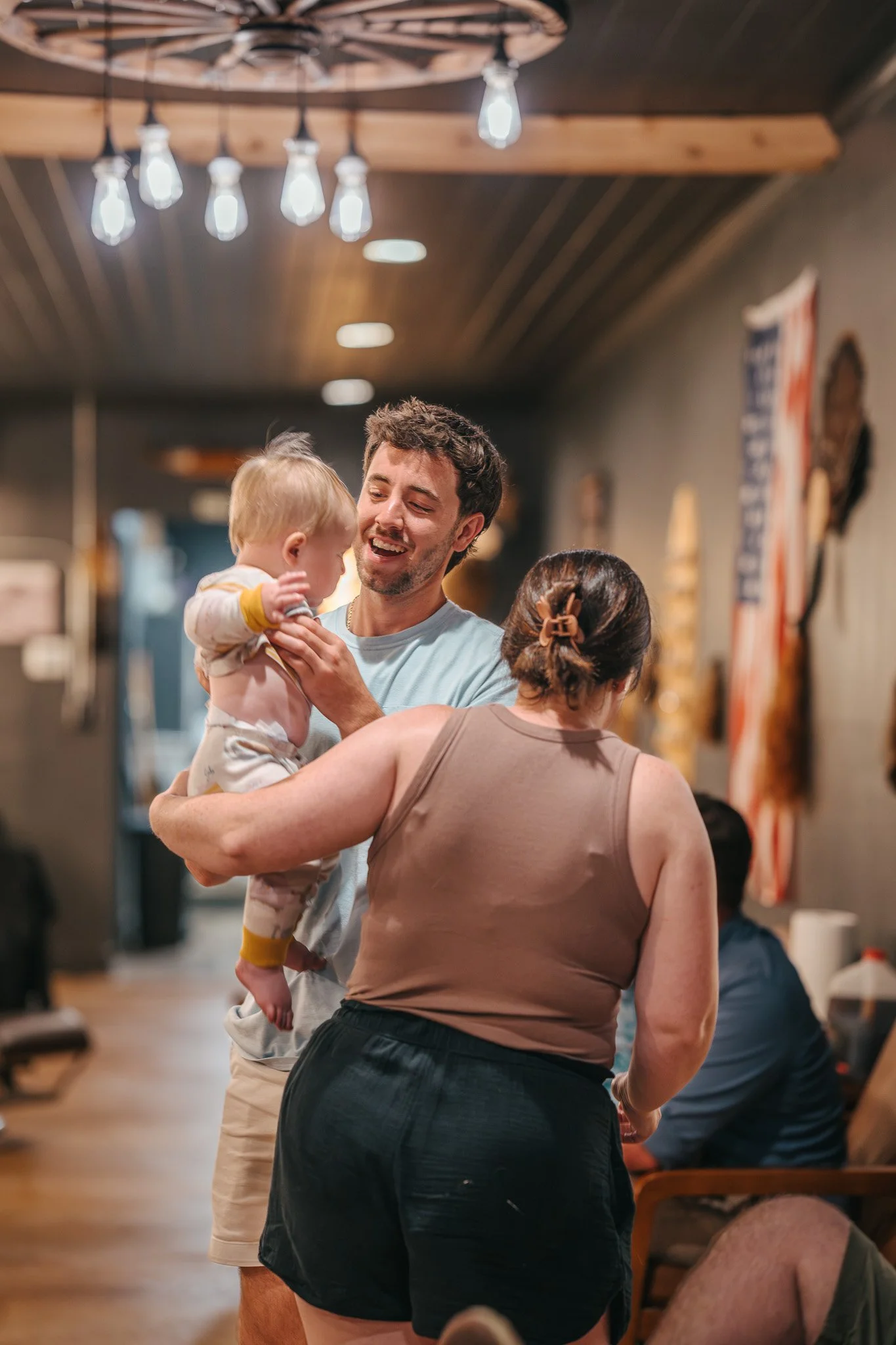 A man holding a toddler in a restaurant or cafe, greeted by a woman with her back to the camera.