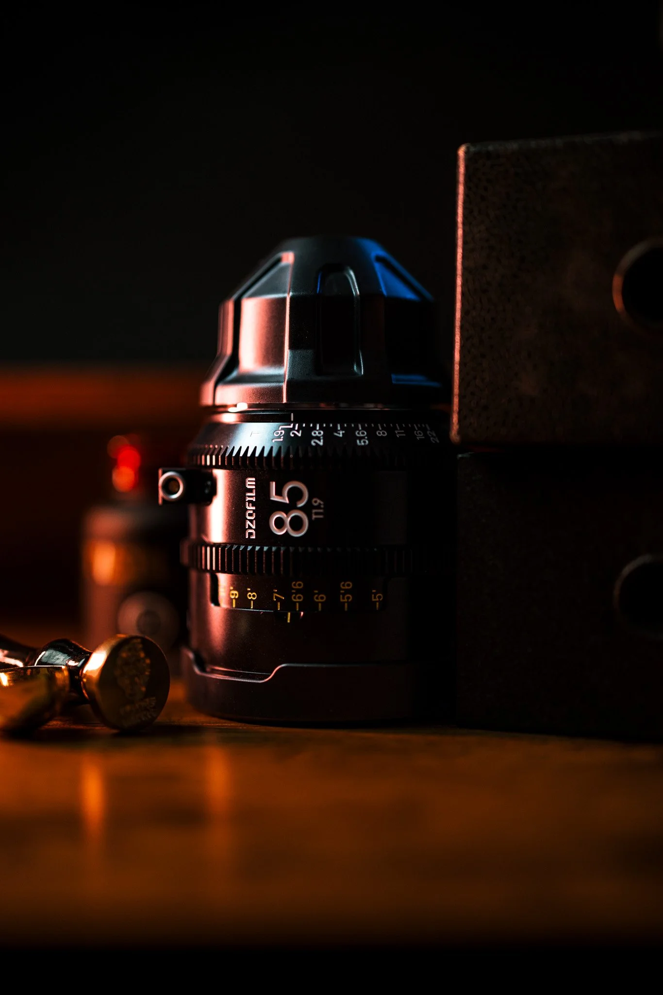 Close-up of a camera lens with a focus on the aperture ring, with a microphone and a textured surface visible nearby, in a dark setting with warm and blue lighting.