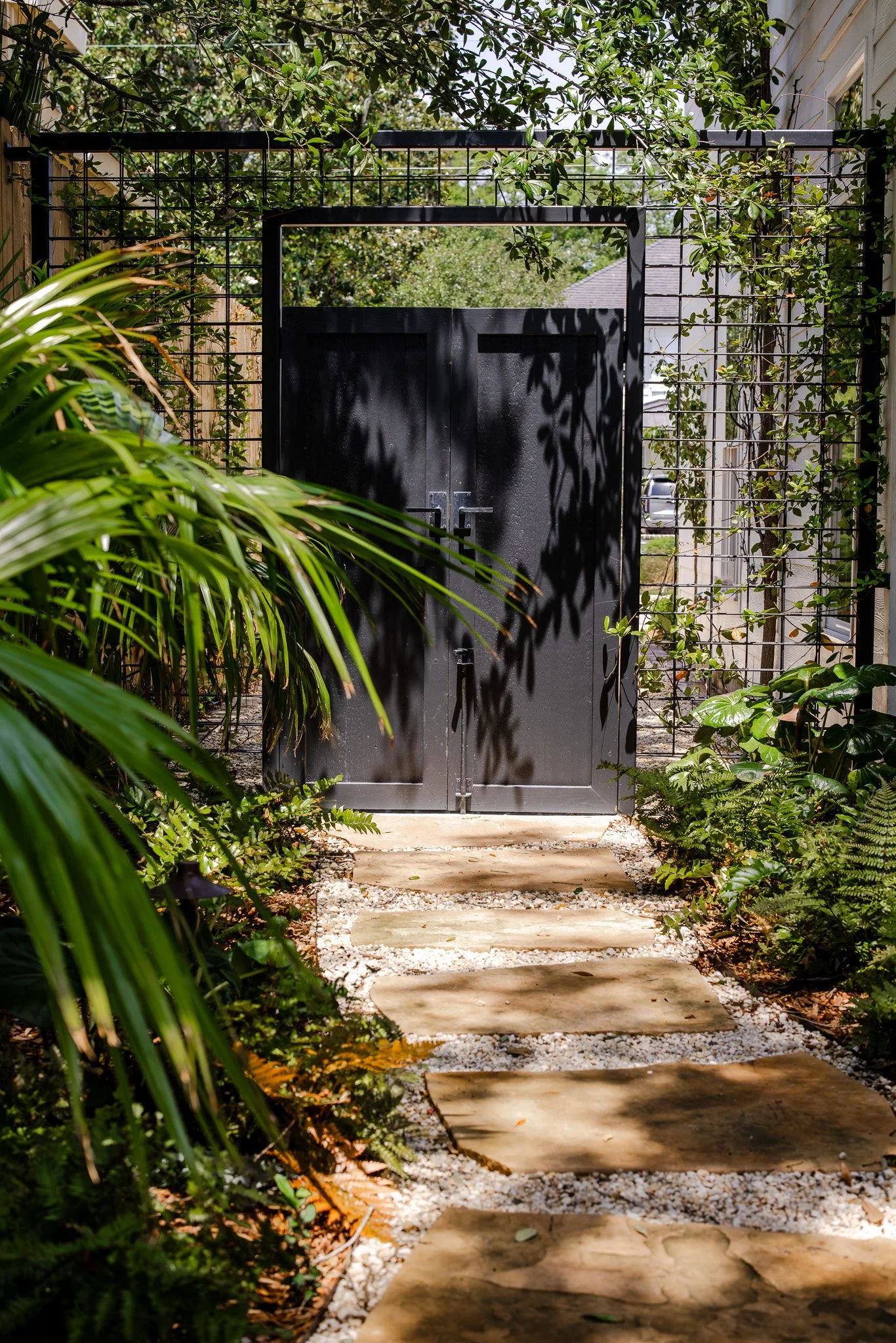 A black metal gate with planters on either side, leading to a garden pathway bordered by lush green foliage and plants, with a white house visible on the right.