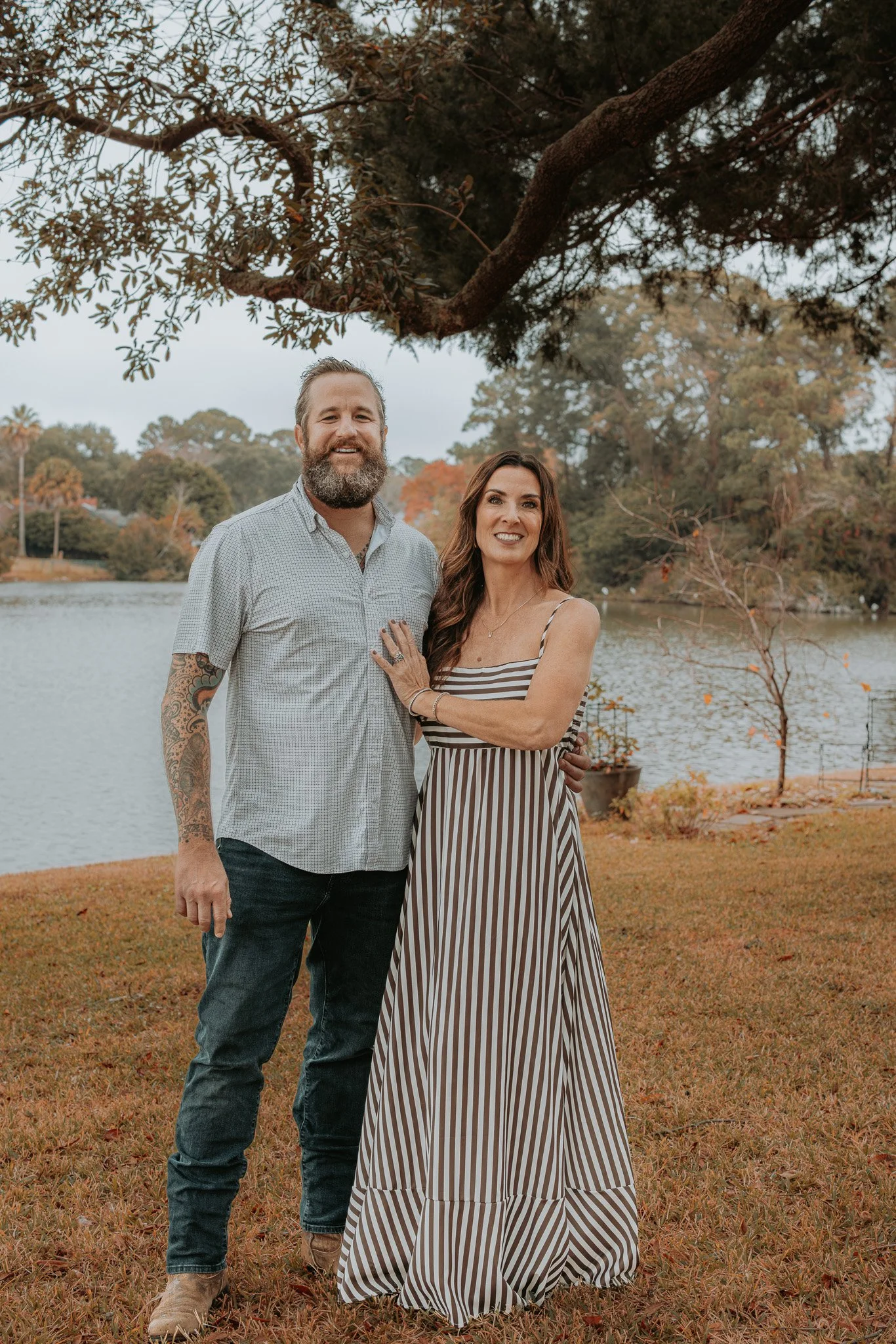 A man and woman standing together outdoors by a lake, under a large tree, smiling at the camera; the woman is wearing a striped dress, and the man is wearing a short-sleeved shirt and jeans.