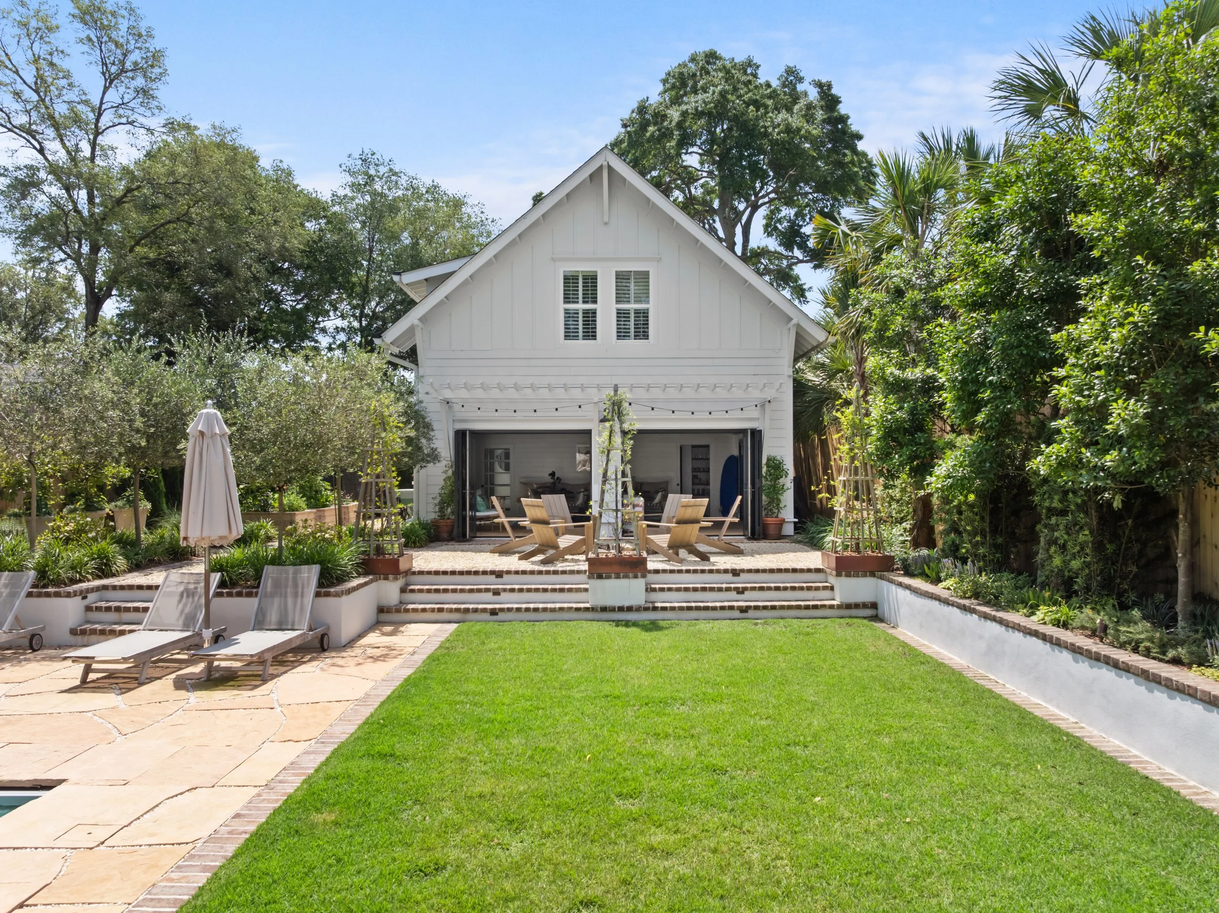 Backyard with white garage, outdoor seating area, green lawn, stone patio, and surrounding trees and bushes.