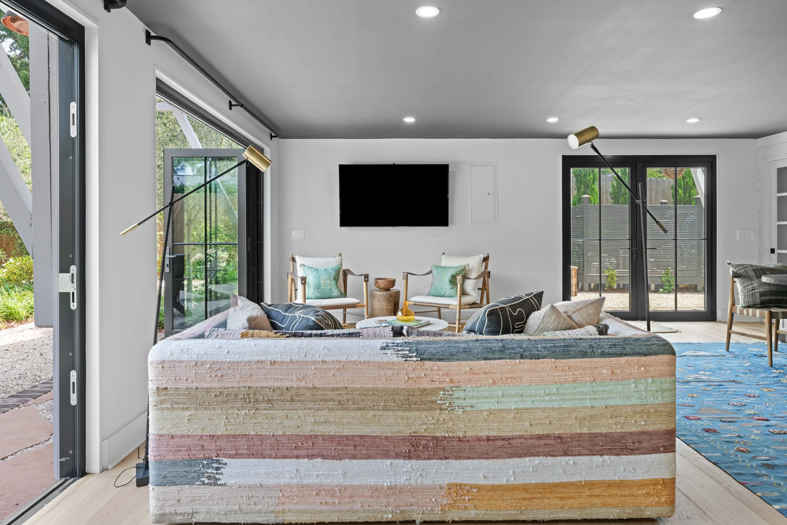 Living room with large pastel-colored striped sofa, two armchairs with blue cushions, a wall-mounted television, and large sliding glass doors opening to a garden.