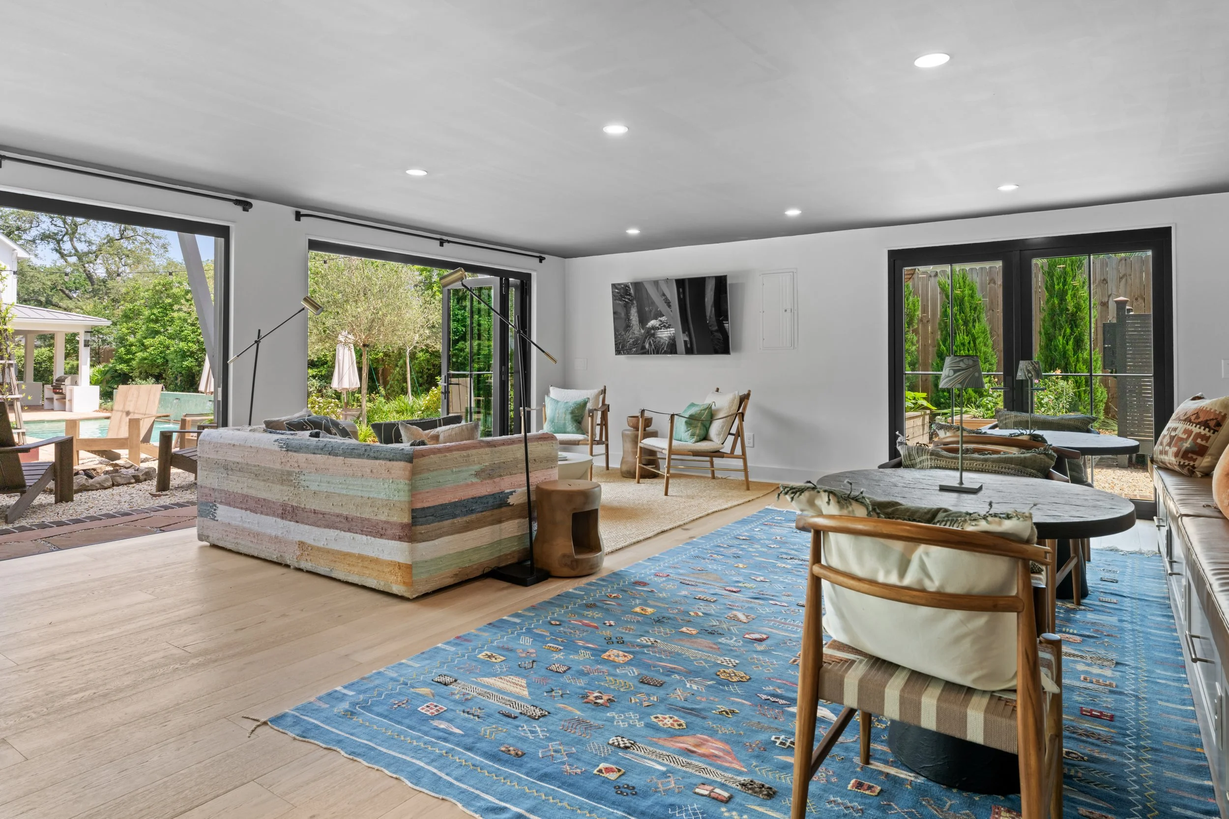Living room with large windows and sliding doors showing outdoor patio and garden, modern furniture including a colorful striped sofa, wooden chairs with cushions, blue patterned rug, and a wall-mounted flat-screen TV.