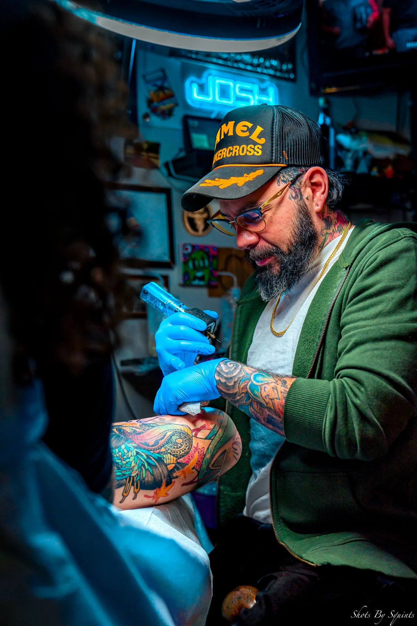 A man with tattoos on his arm getting a tattoo on his thigh from an artist wearing blue gloves. The scene is illuminated by blue neon lights, with the word 'JOSH' visible in the background.