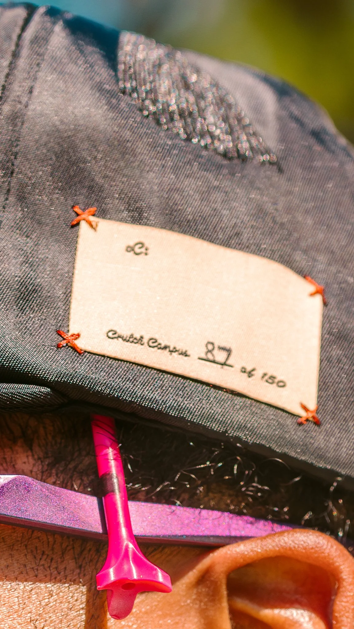 Close-up of a clothing label sewn onto dark gray fabric, with a pink safety pin and part of a brown ear visible in the foreground.