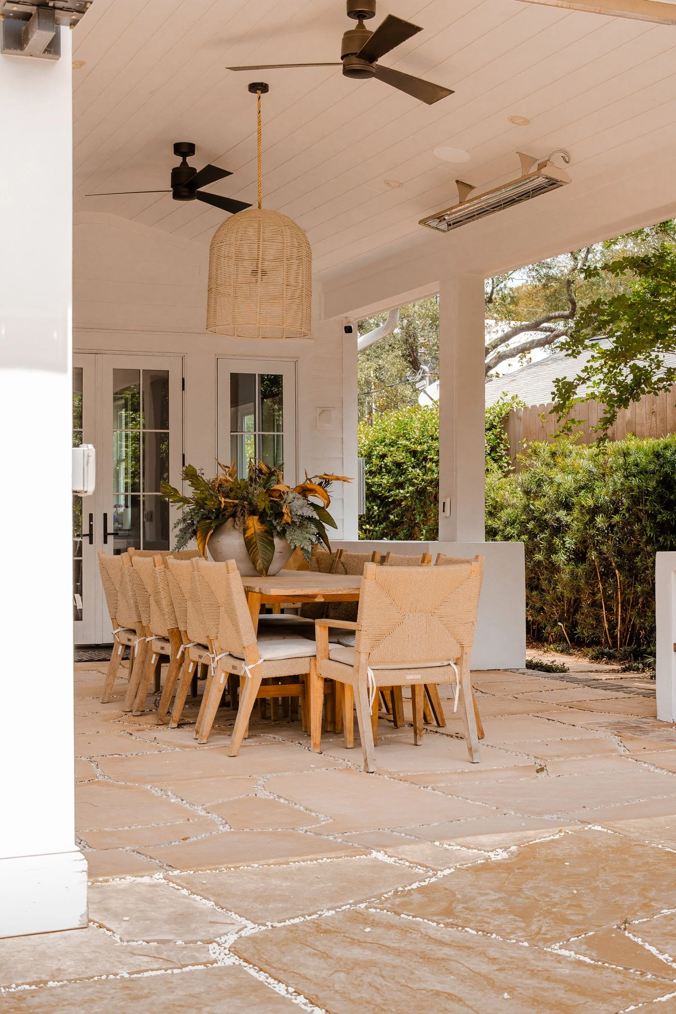 Outdoor patio with a wooden dining table, wicker chairs, a potted plant centerpiece, ceiling fans, an outdoor heater, and a view of trees and a wooden fence.